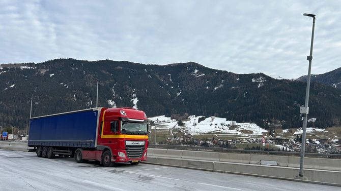 Een rode vrachtwagen met een blauwe oplegger rijdt over een besneeuwde weg met bergen op de achtergrond.