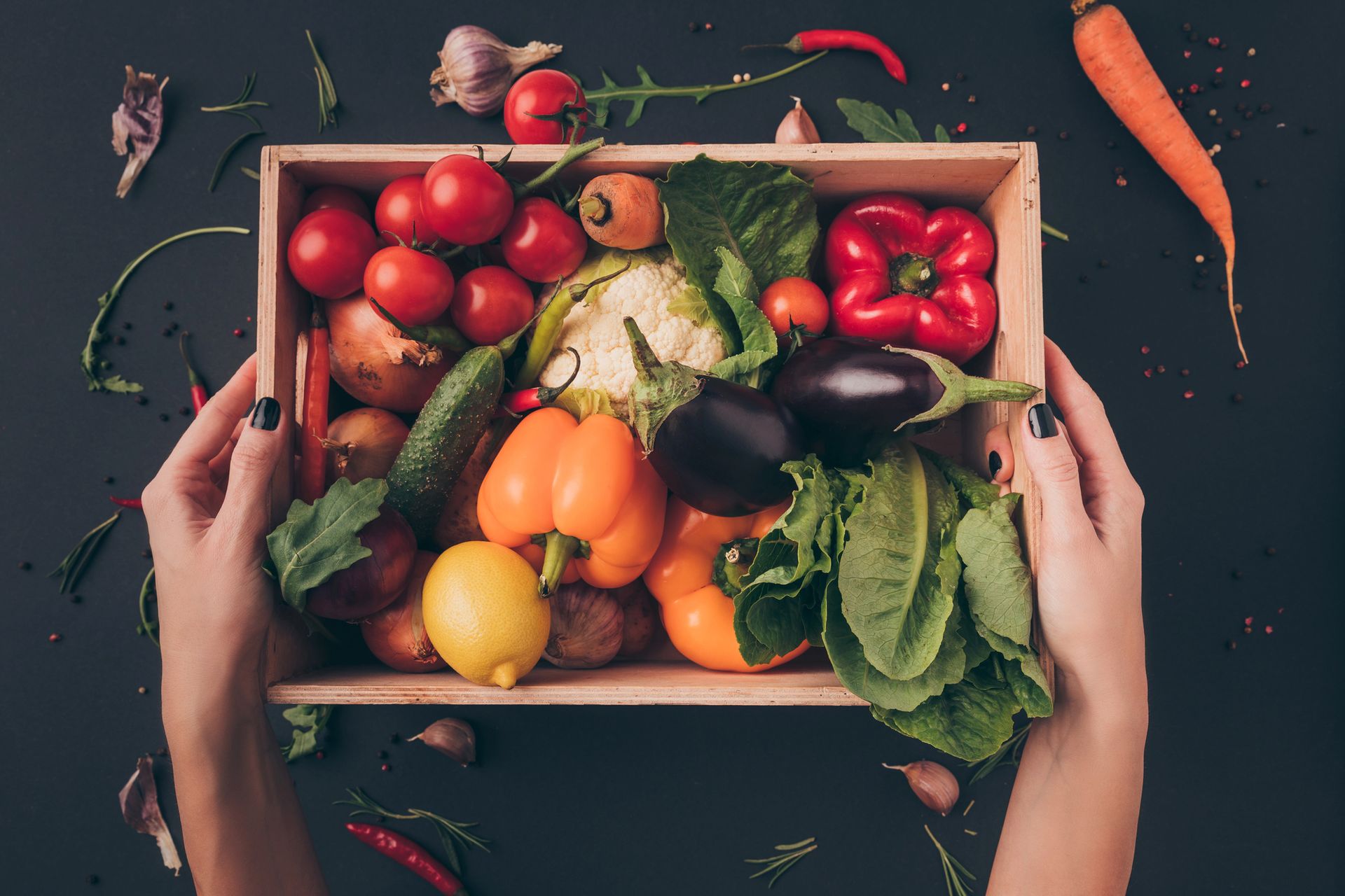 Mani che tengono una cassa di legno piena di verdure colorate: pomodori, peperoni, cetrioli, lattuga e molto altro.