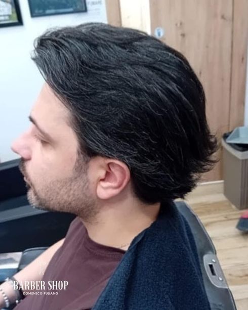 Profilo laterale di un uomo con capelli e barba scuri pettinati all'indietro, seduto su una poltrona da parrucchiere, prima di un taglio di capelli.