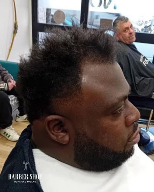 Uomo con capelli afro e barba rasati, seduto sulla poltrona di un barbiere, ritratto di profilo.