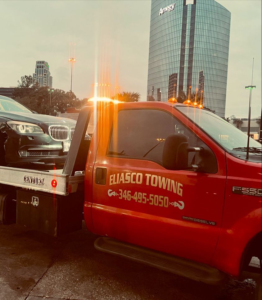 Red Elasi towing truck beside a disabled car at dusk with city buildings in the background