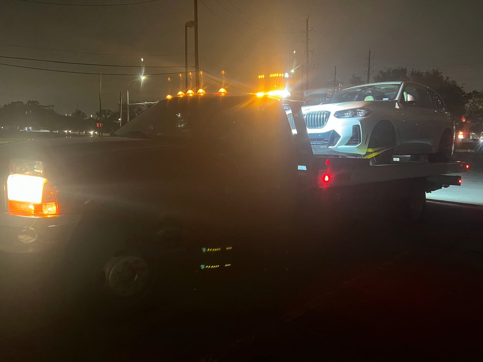 Tow truck loading a white SUV at night on a wet street, with bright lights and traffic in the background
