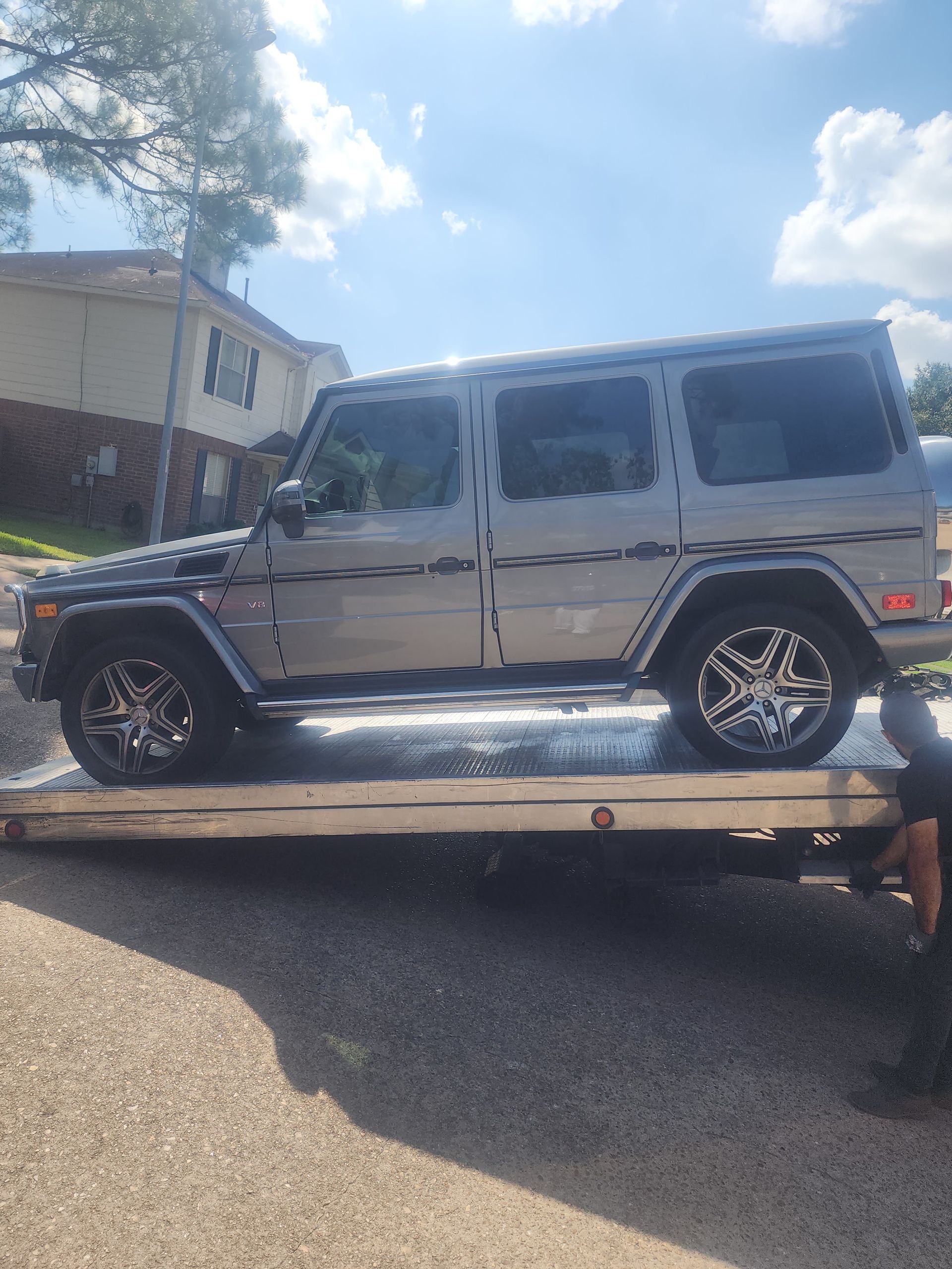 Silver SUV on a flatbed tow truck in bright sunlight, with a building and trees in the background