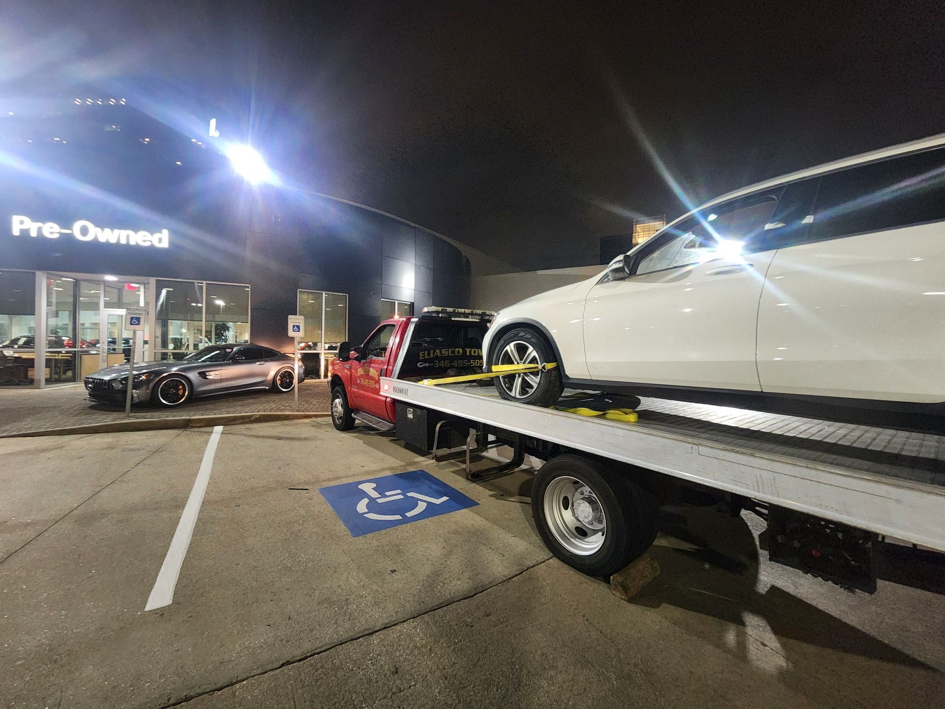 Tow truck loading a white car at night outside a Prenatal store; red car parked nearby.