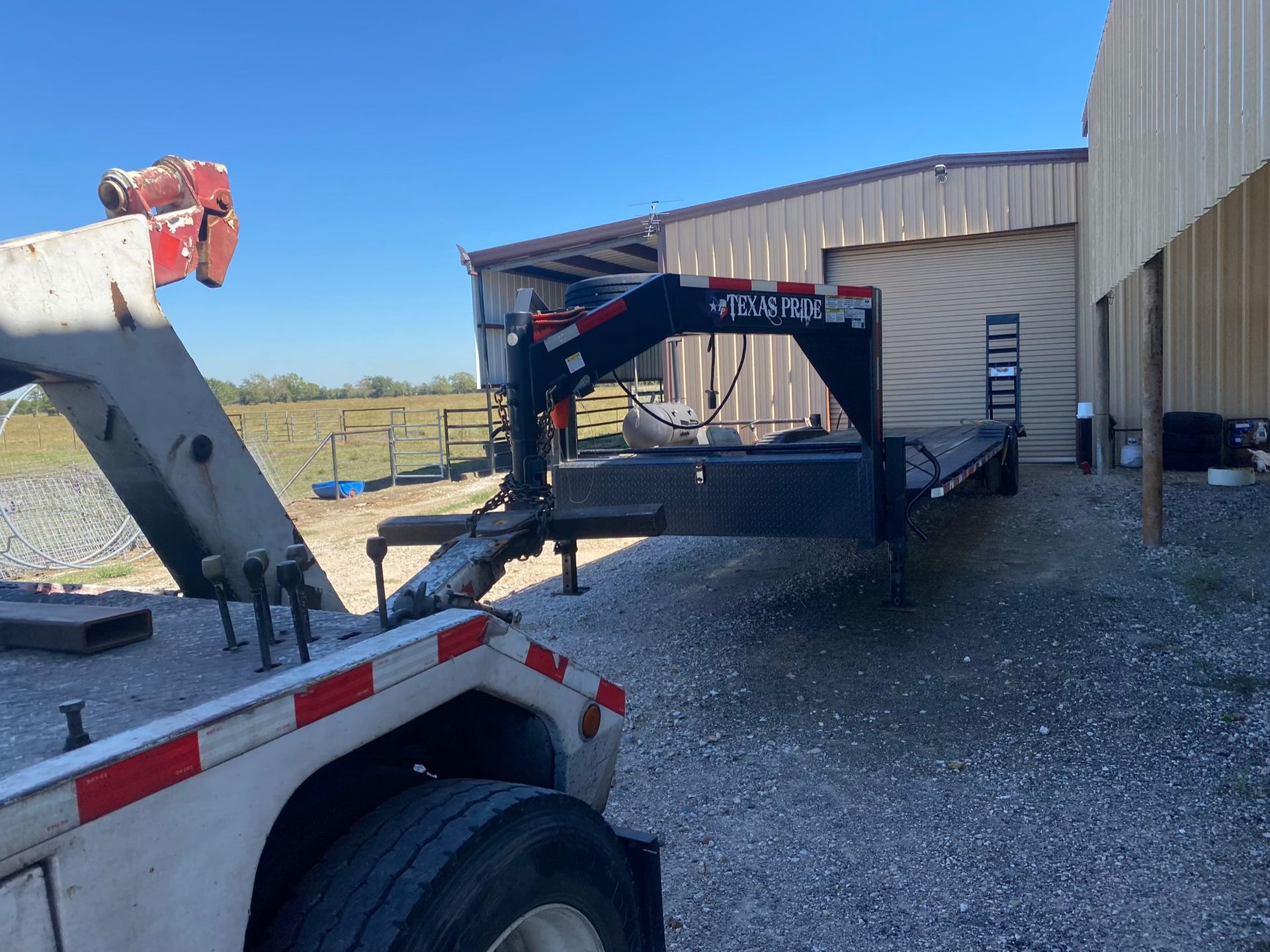 Black gooseneck trailer backed into a tan metal barn beside a gravel yard