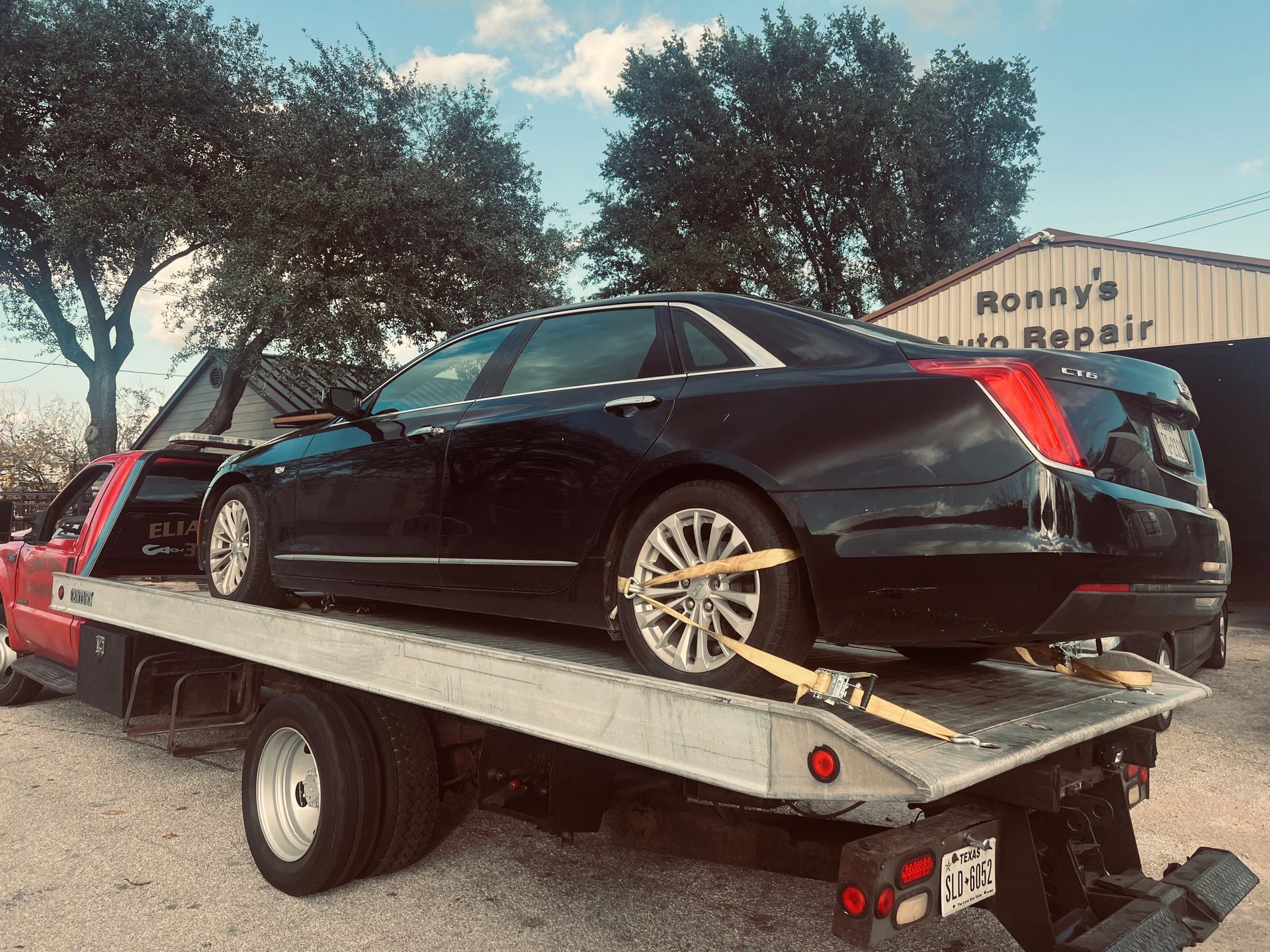 Black sedan on a tow truck outside a repair shop, with trees and blue sky in the background