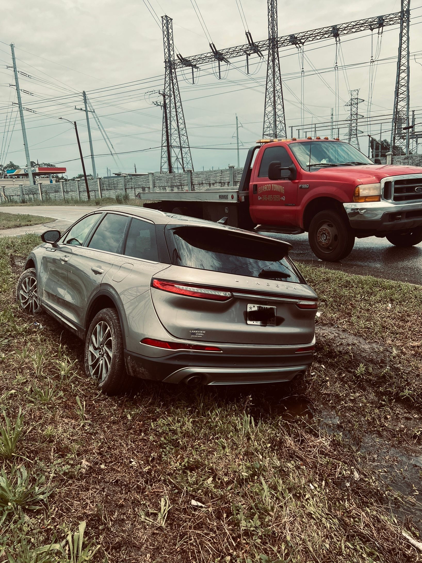 Silver sedan stopped on a muddy roadside near a red tow truck under power lines