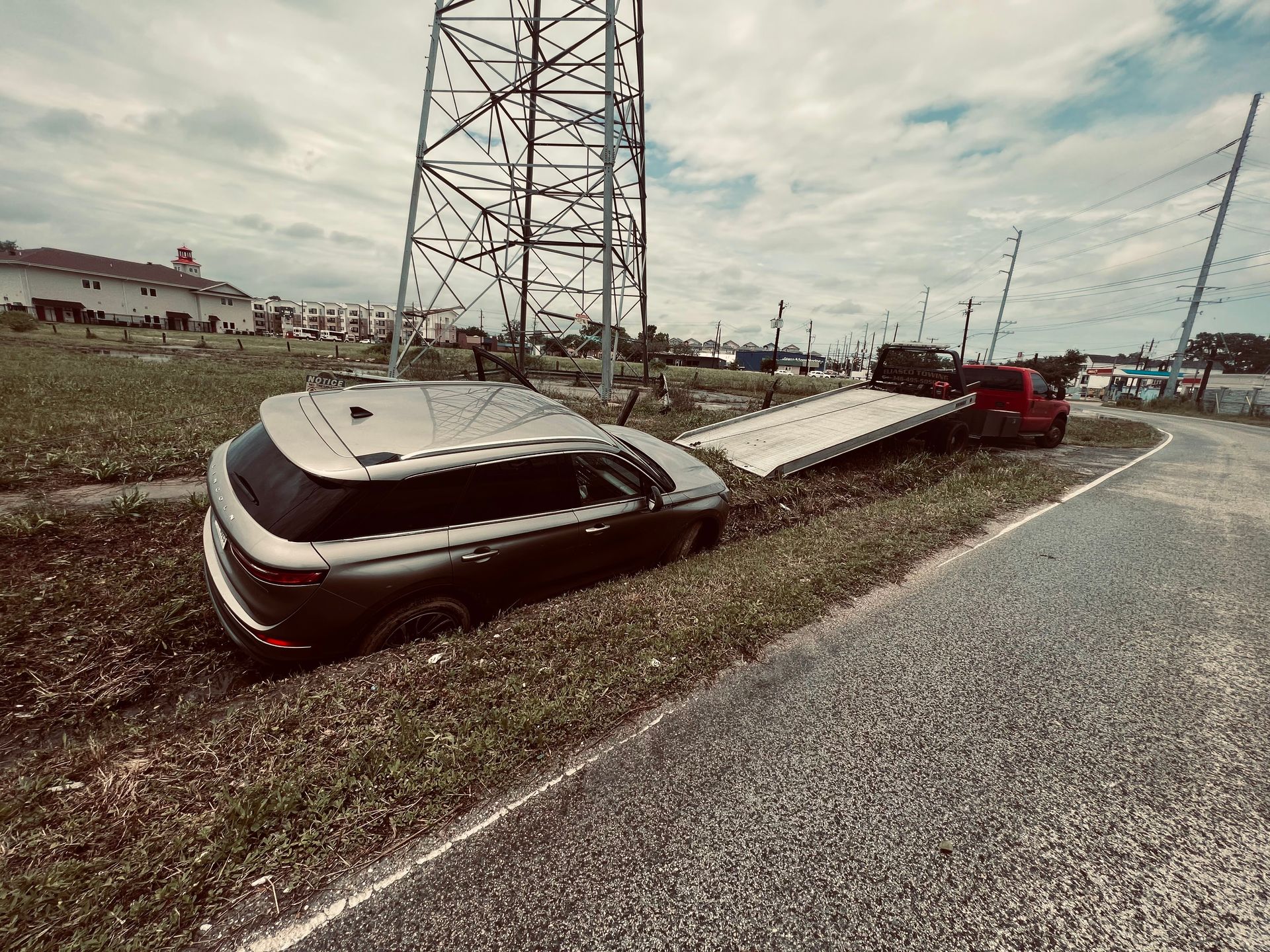 A black car overturned in a grassy ditch beside a road, with a tow truck nearby under power lines.