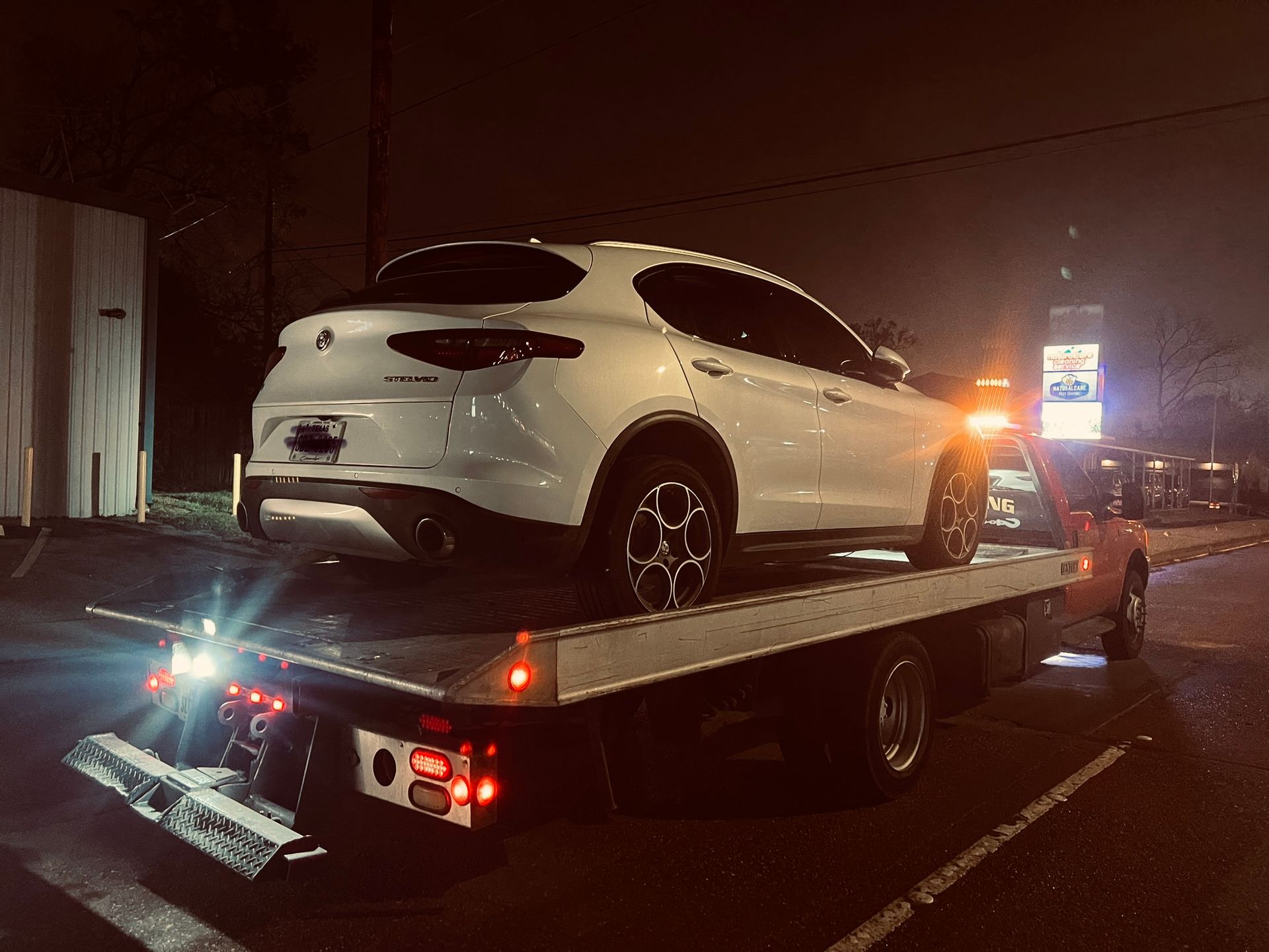 White SUV on a flatbed tow truck at night on a street, with flashing lights.