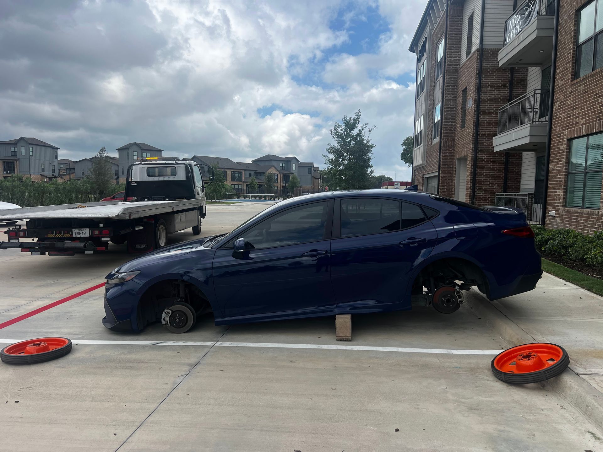 Blue sedan with flat tires on a driveway, near a tow truck and apartment building.