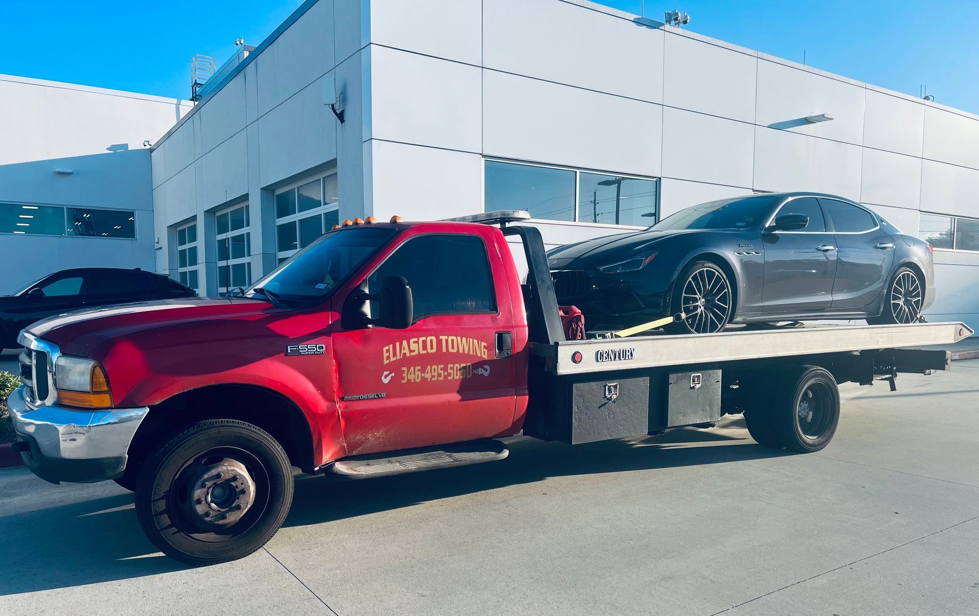 Red flatbed tow truck carrying a gray sedan outside an auto service building