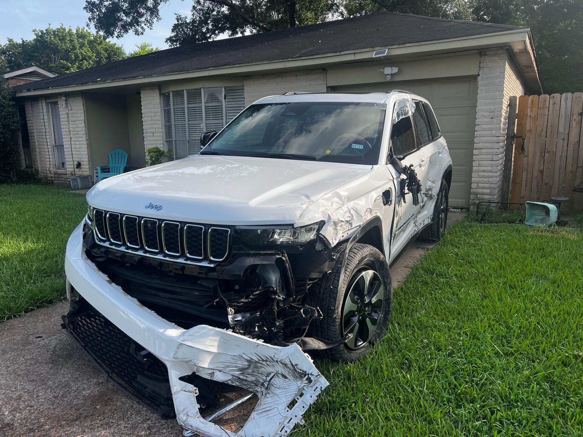 Damaged white SUV with front bumper torn off, parked in a driveway in front of a house.