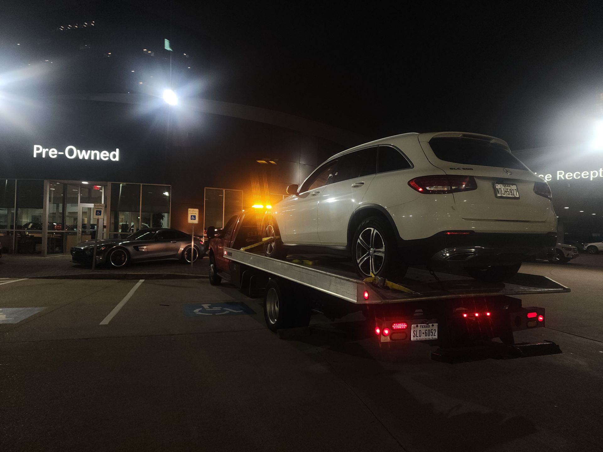 White SUV on a flatbed tow truck at night outside a pre-owned car dealership.