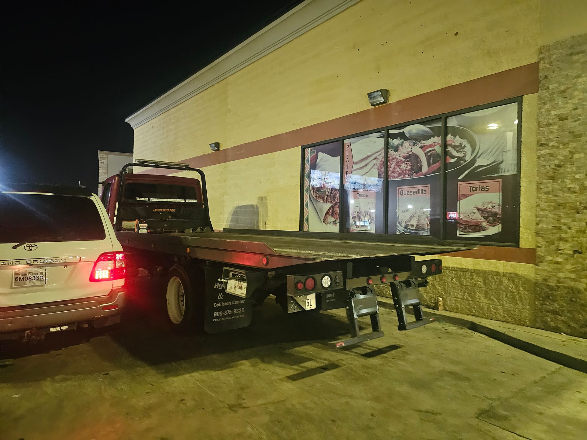 Tow truck backing into a storefront parking spot beside a white car at night
