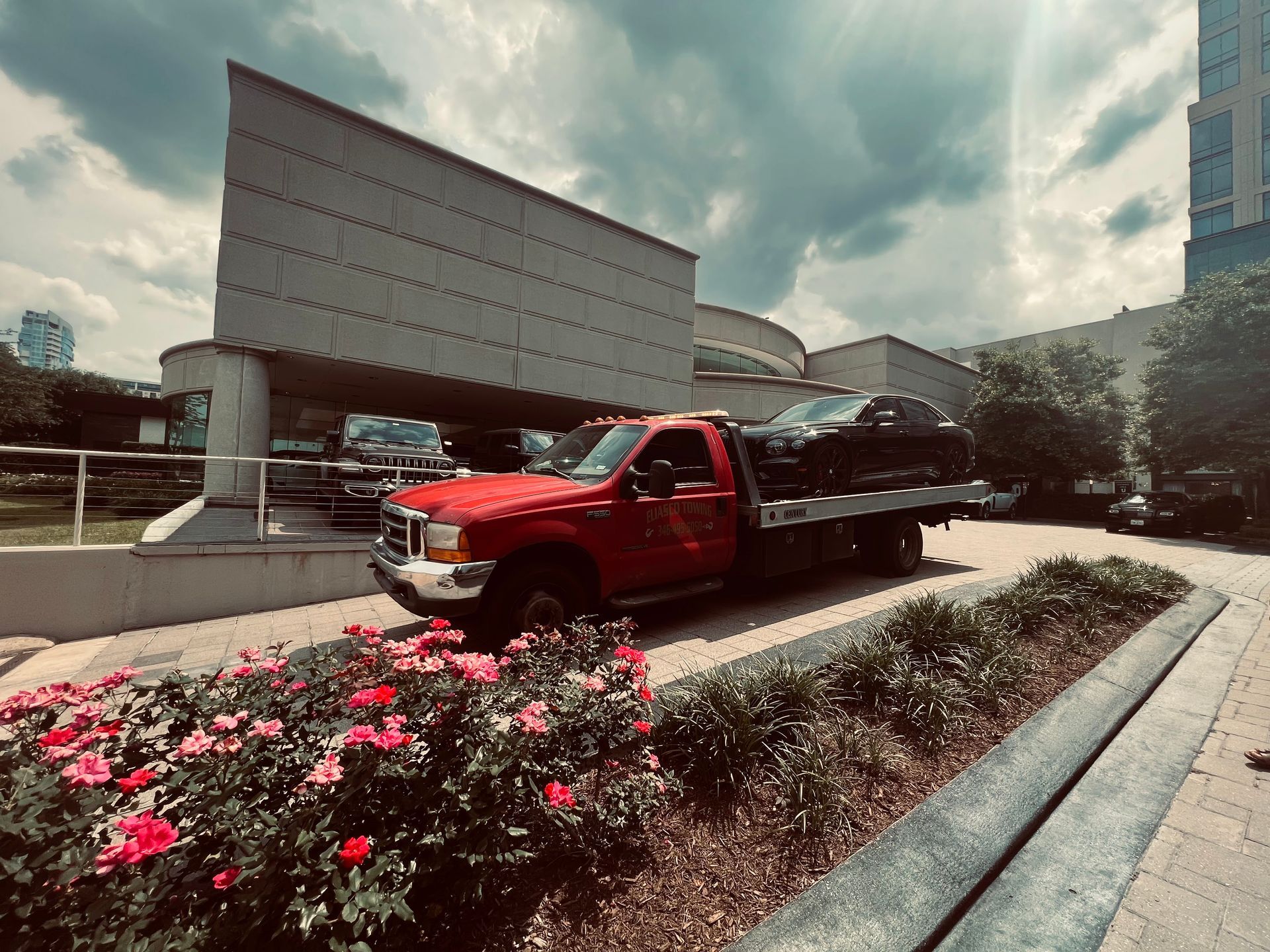 Red flatbed truck on a city street beside pink flowers and modern buildings under a cloudy sky