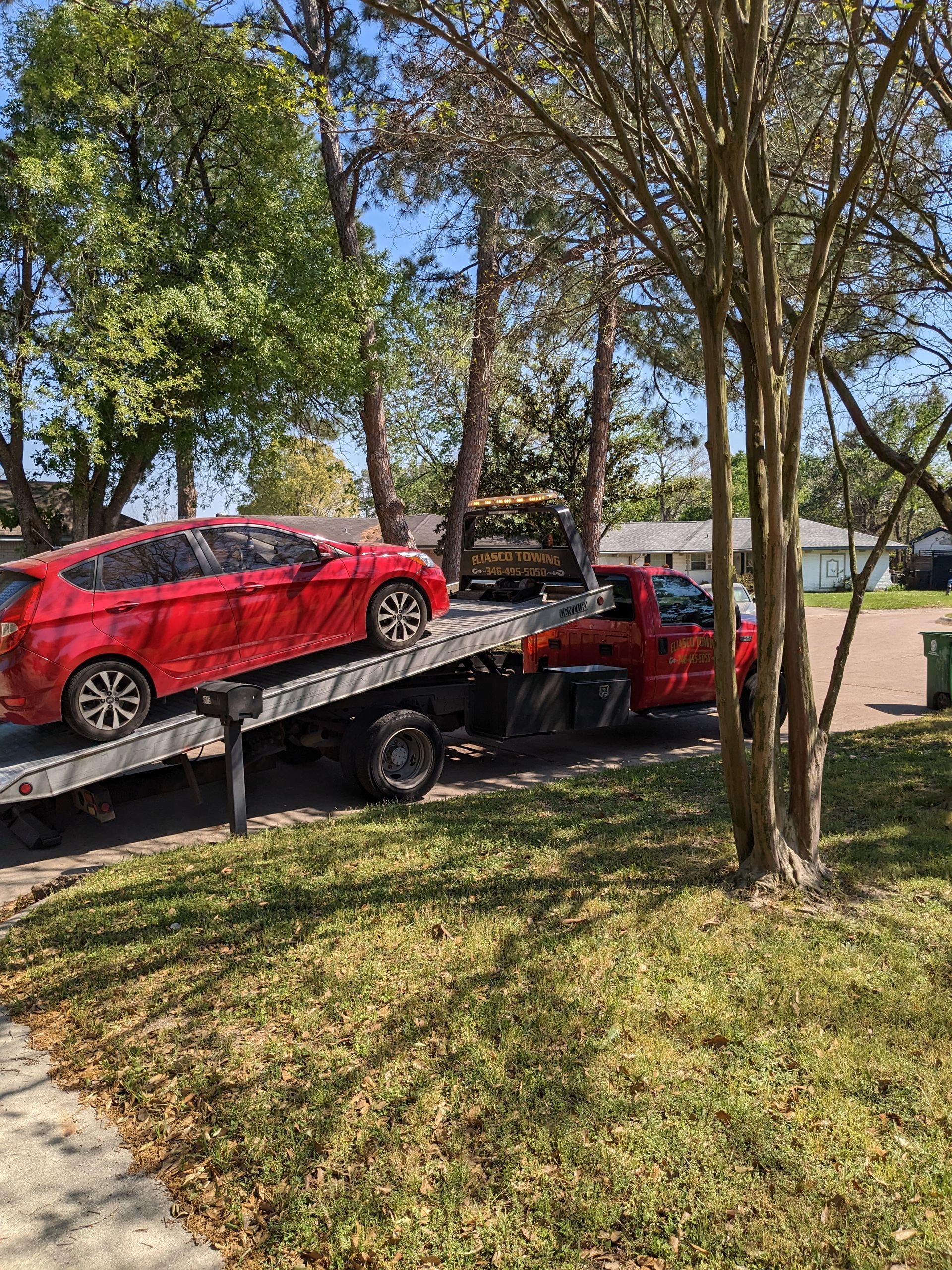 Red sedan on a tow truck in a sunny residential yard with trees and a house in the background