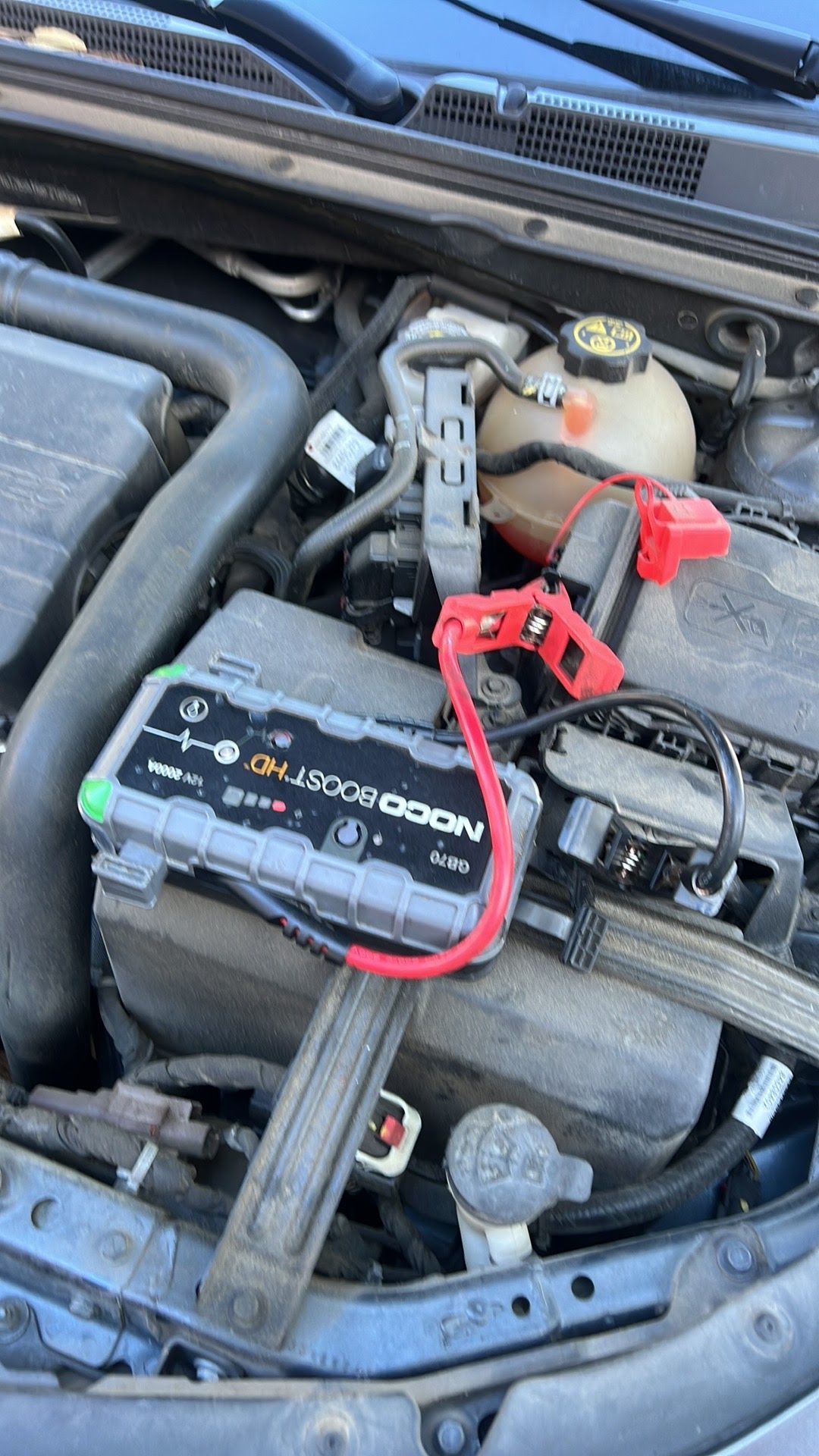 Car battery in engine bay with red jumper cable attached to the positive terminal.