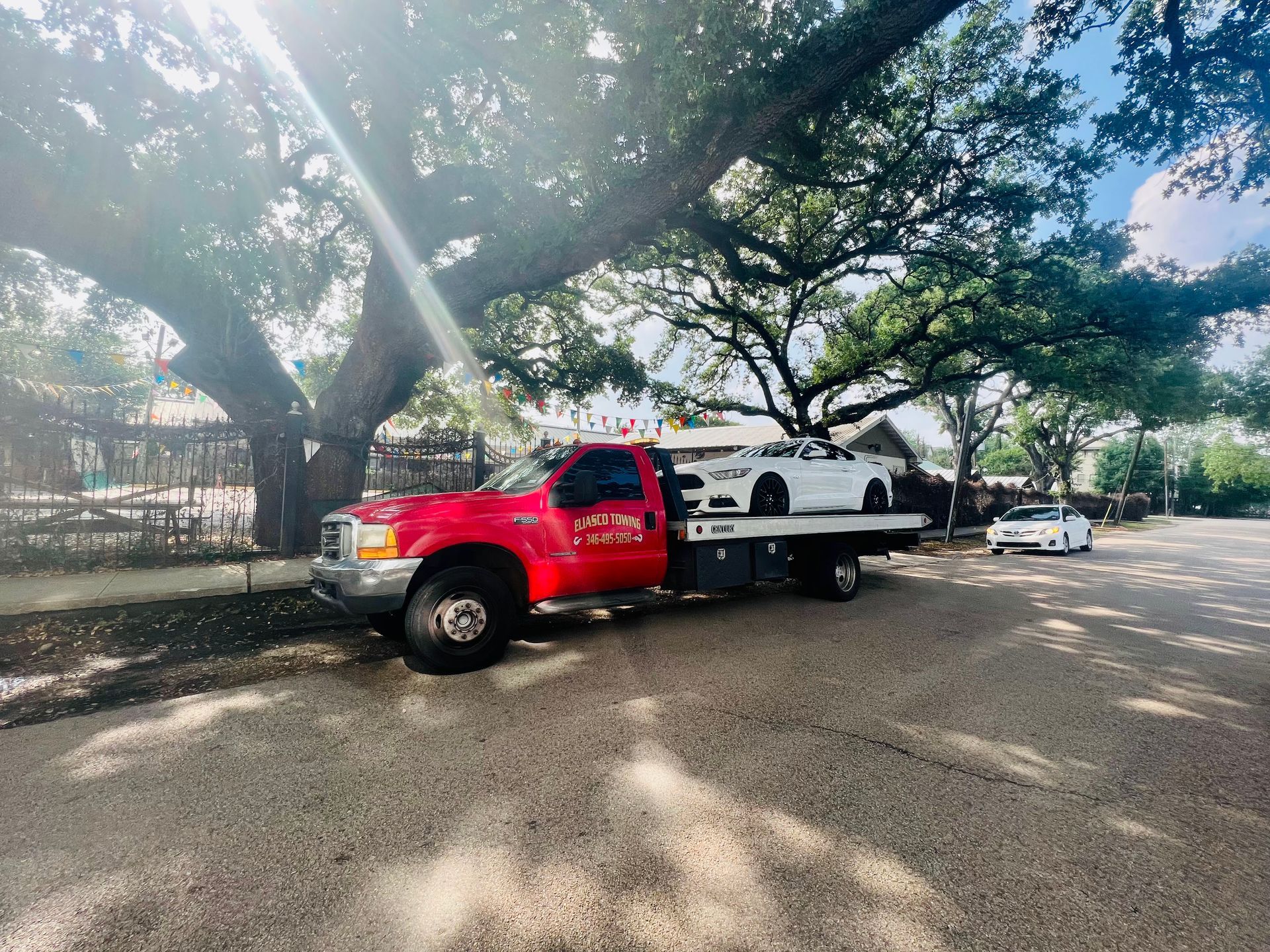 Red tow truck parked on a tree-lined street beside a white car carrier, with sunlight and shadows on the road.