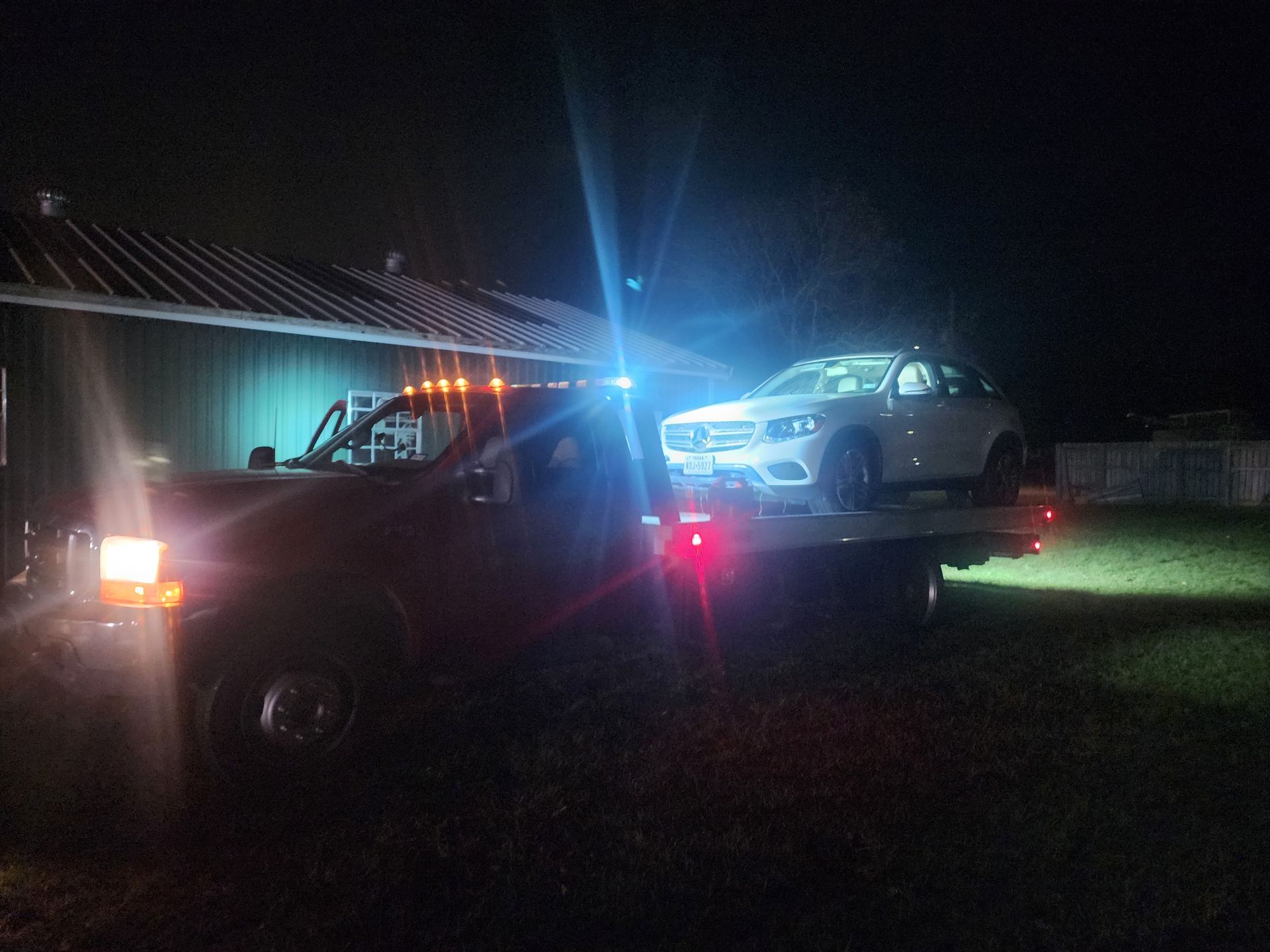 Tow truck at night lifting a silver car, with bright lights and a dark roadside background