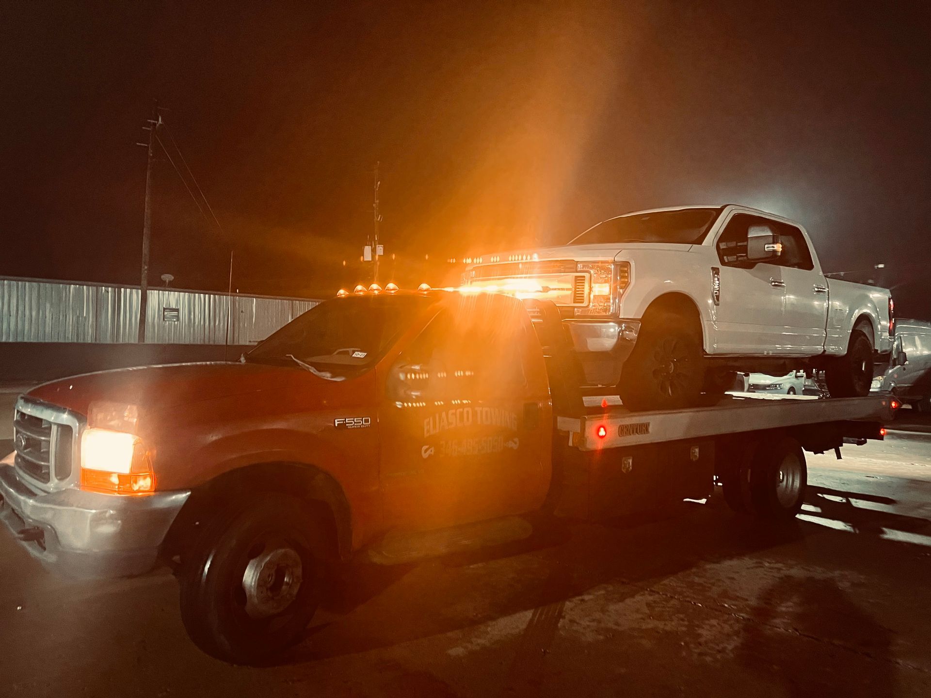 Tow truck at night carrying a white pickup on its flatbed, amber lights flashing.