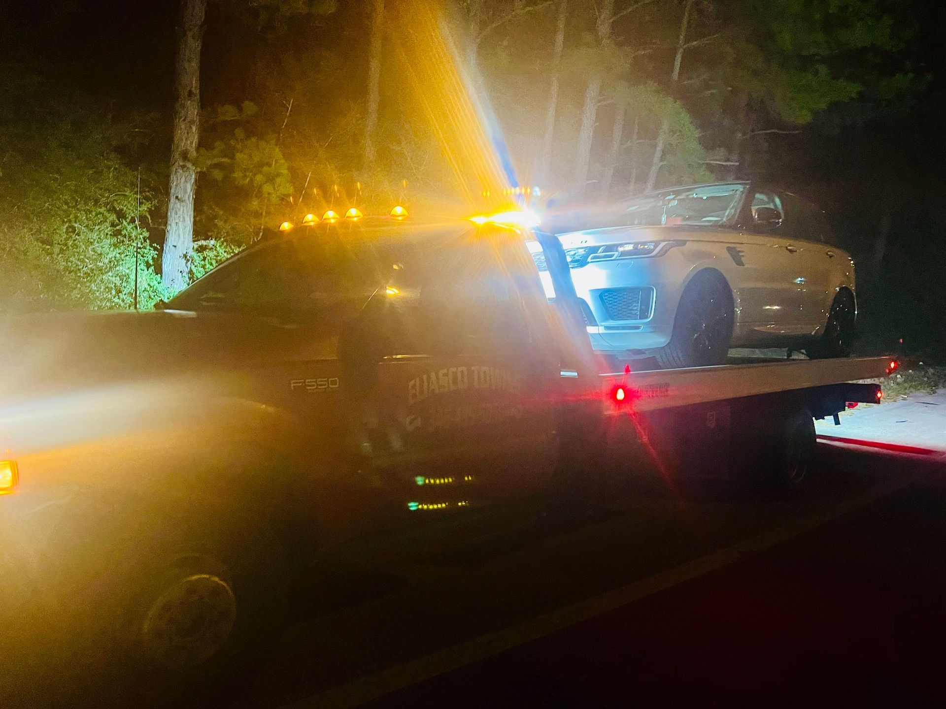 White car on a tow truck at night with flashing lights and glare from headlights