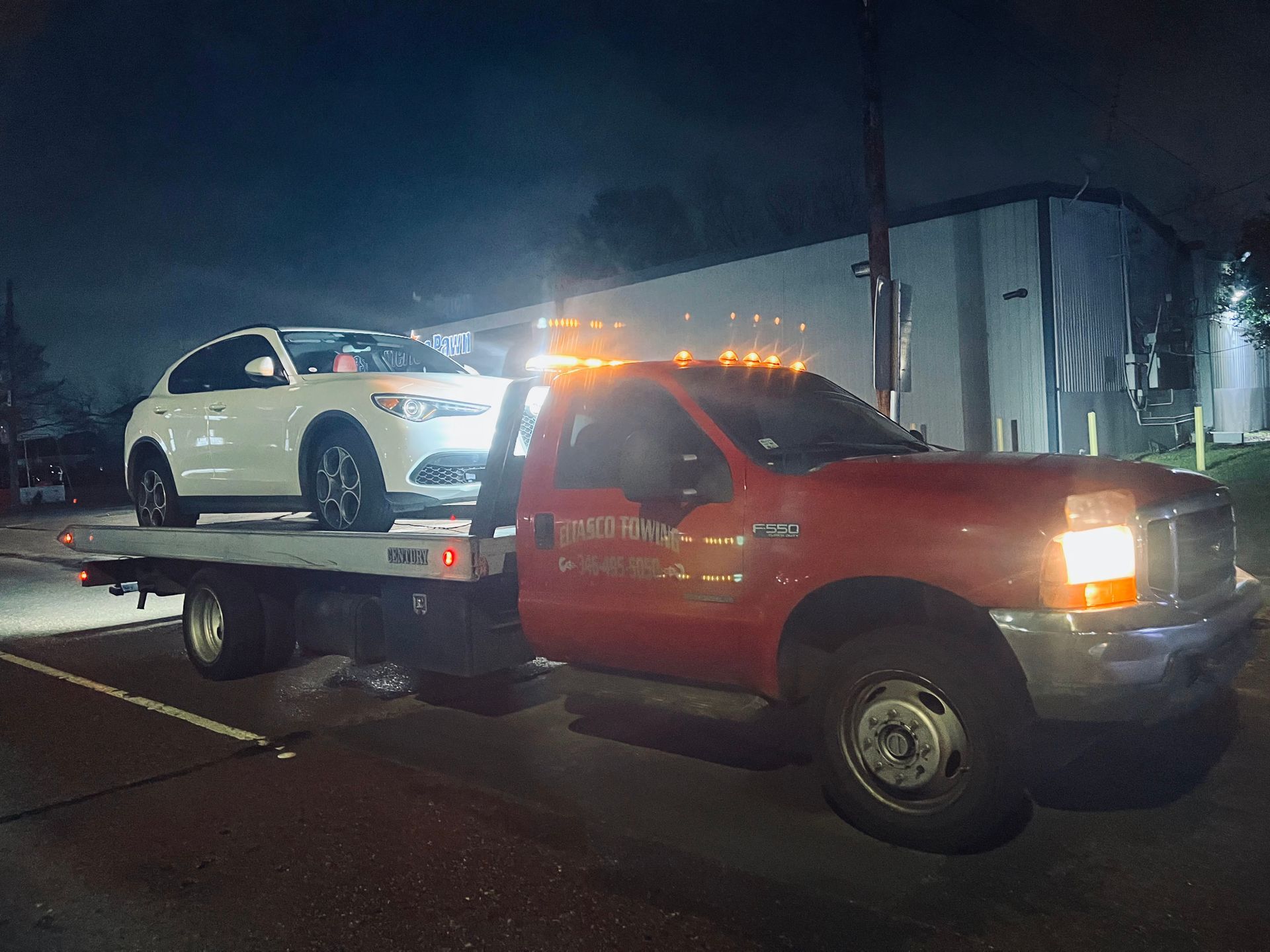 Tow truck carrying a white SUV at night on a wet street, red cab with flashing amber lights