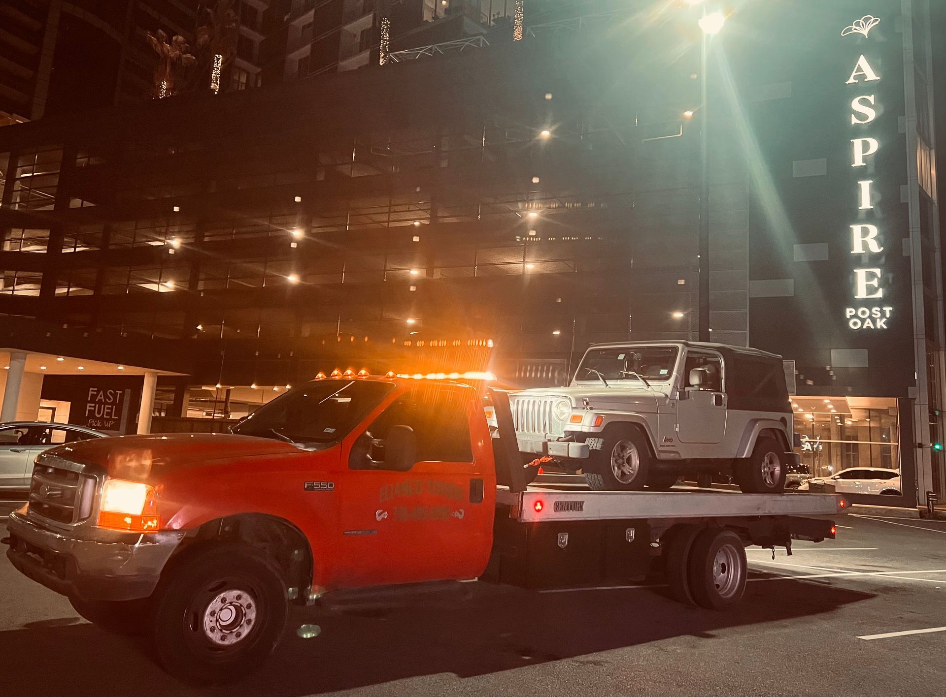 Tow truck hauling a white Jeep at night outside the Aspire building with bright lights