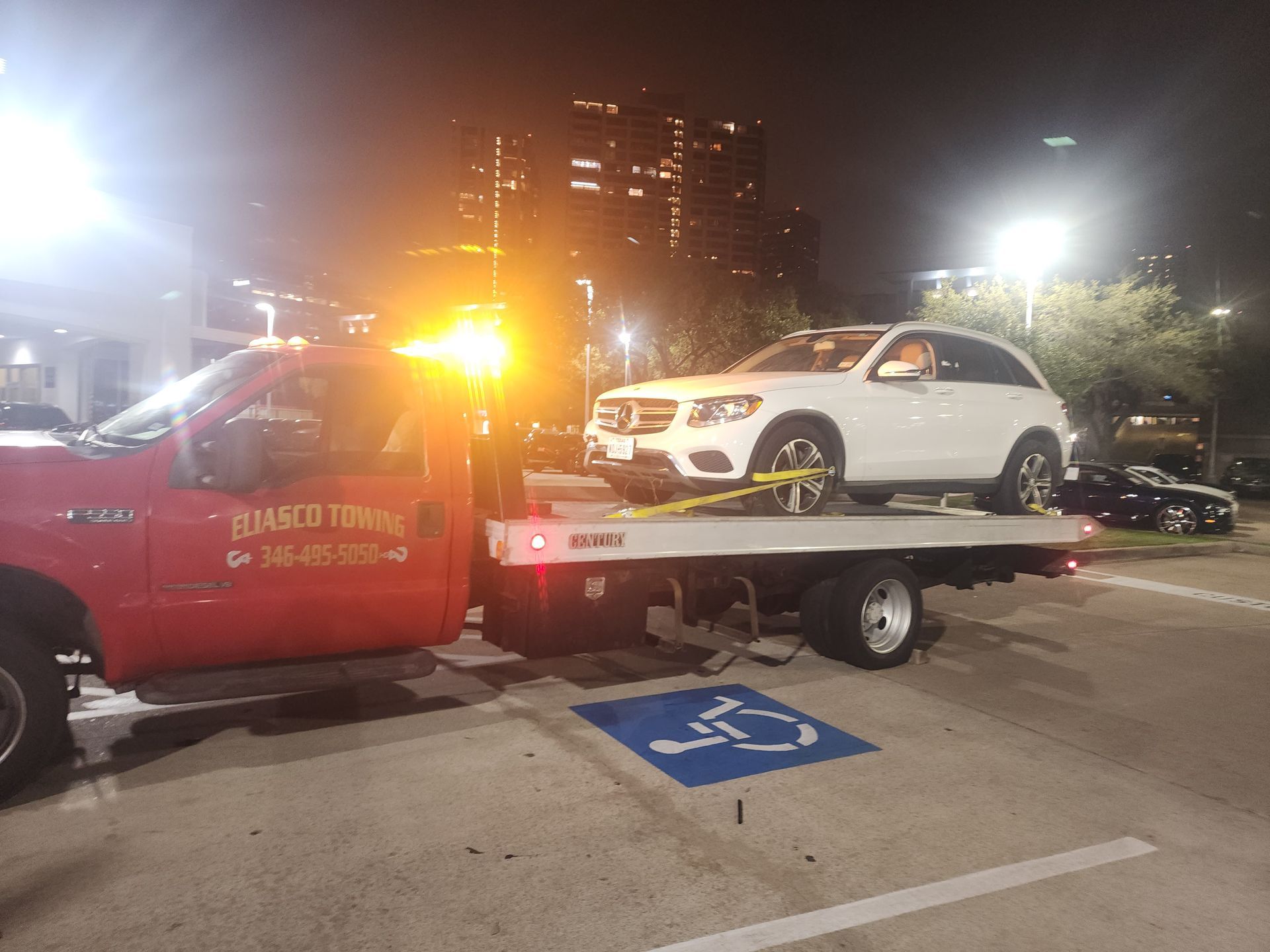 Tow truck hauling a white car in a parking lot at night, with flashing amber lights.