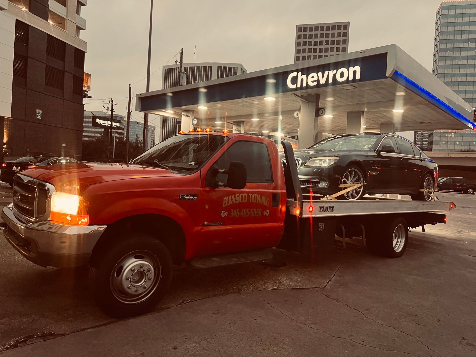 Red tow truck carrying a black car outside a Chevron gas station at dusk
