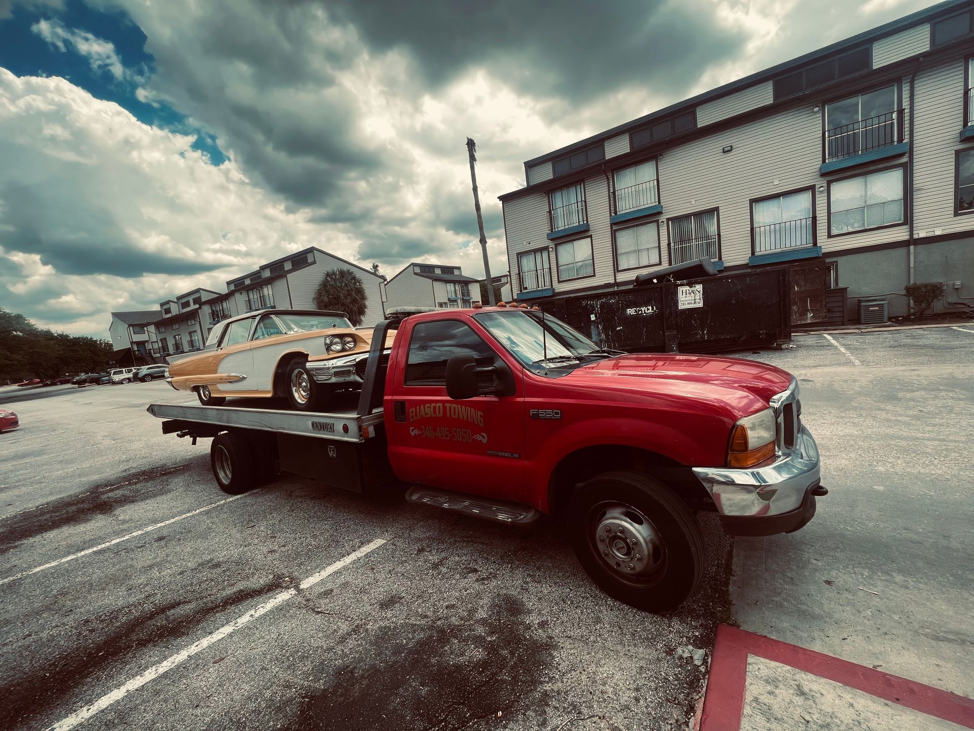 Red tow truck carrying a white sedan in an industrial parking lot under cloudy skies