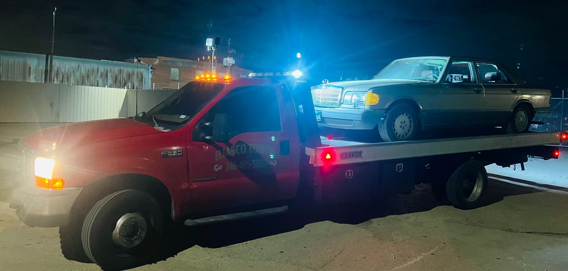 Red tow truck carrying a blue car at night on a street, with flashing emergency lights.