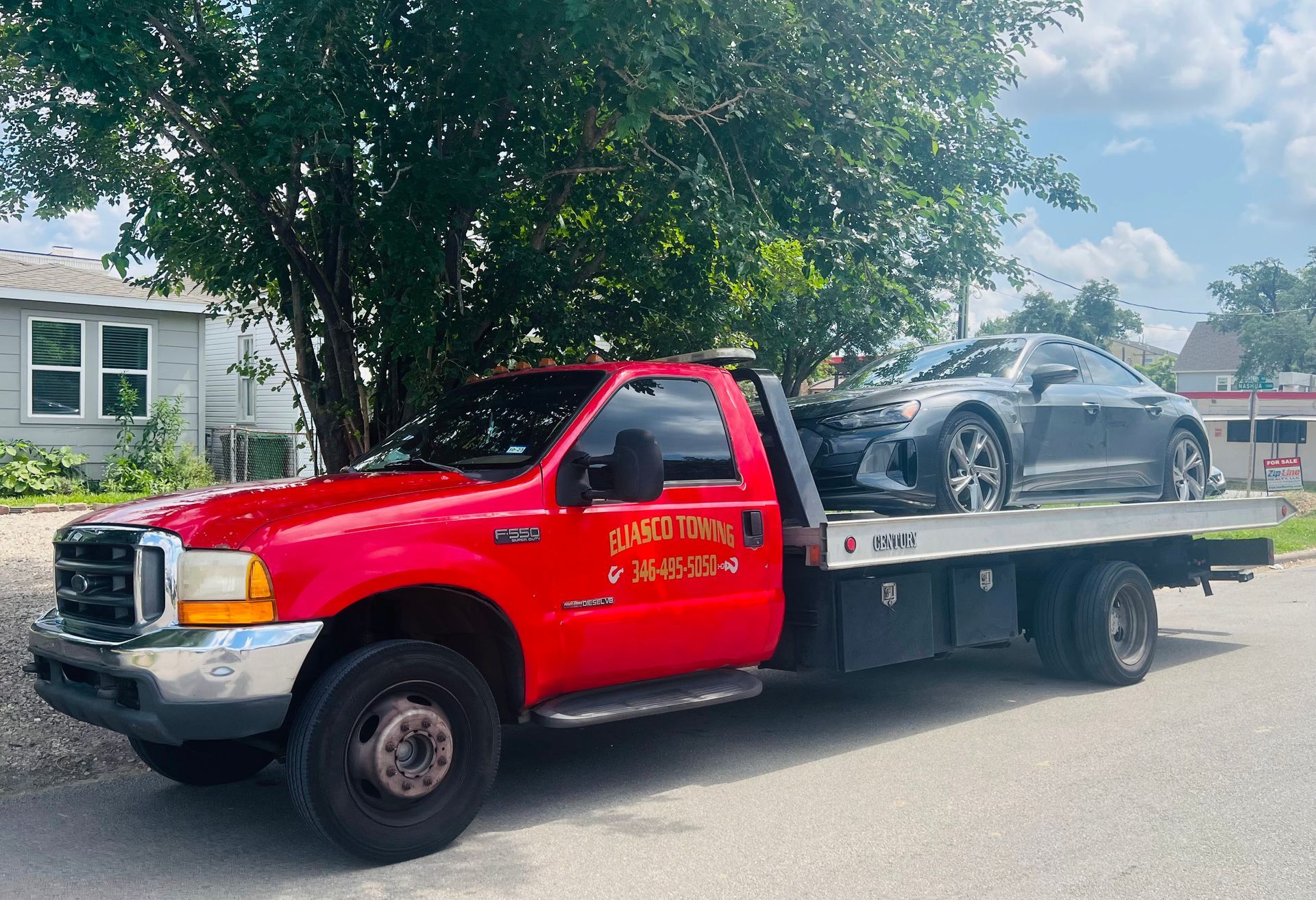 Red tow truck carrying a damaged silver car on a flatbed beside a residential street