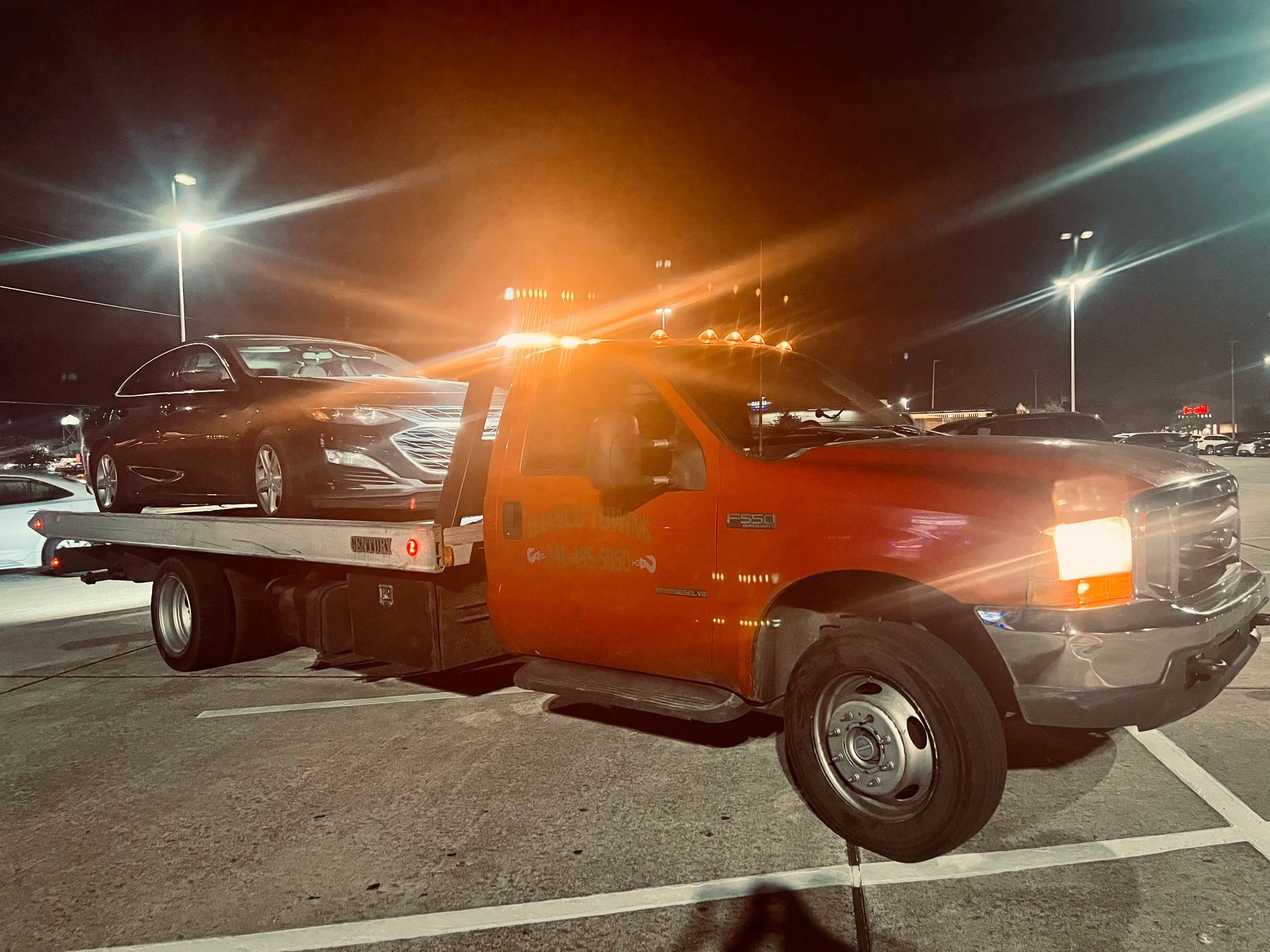 Orange tow truck carrying a silver car at night in a parking lot, with bright lights and cones nearby