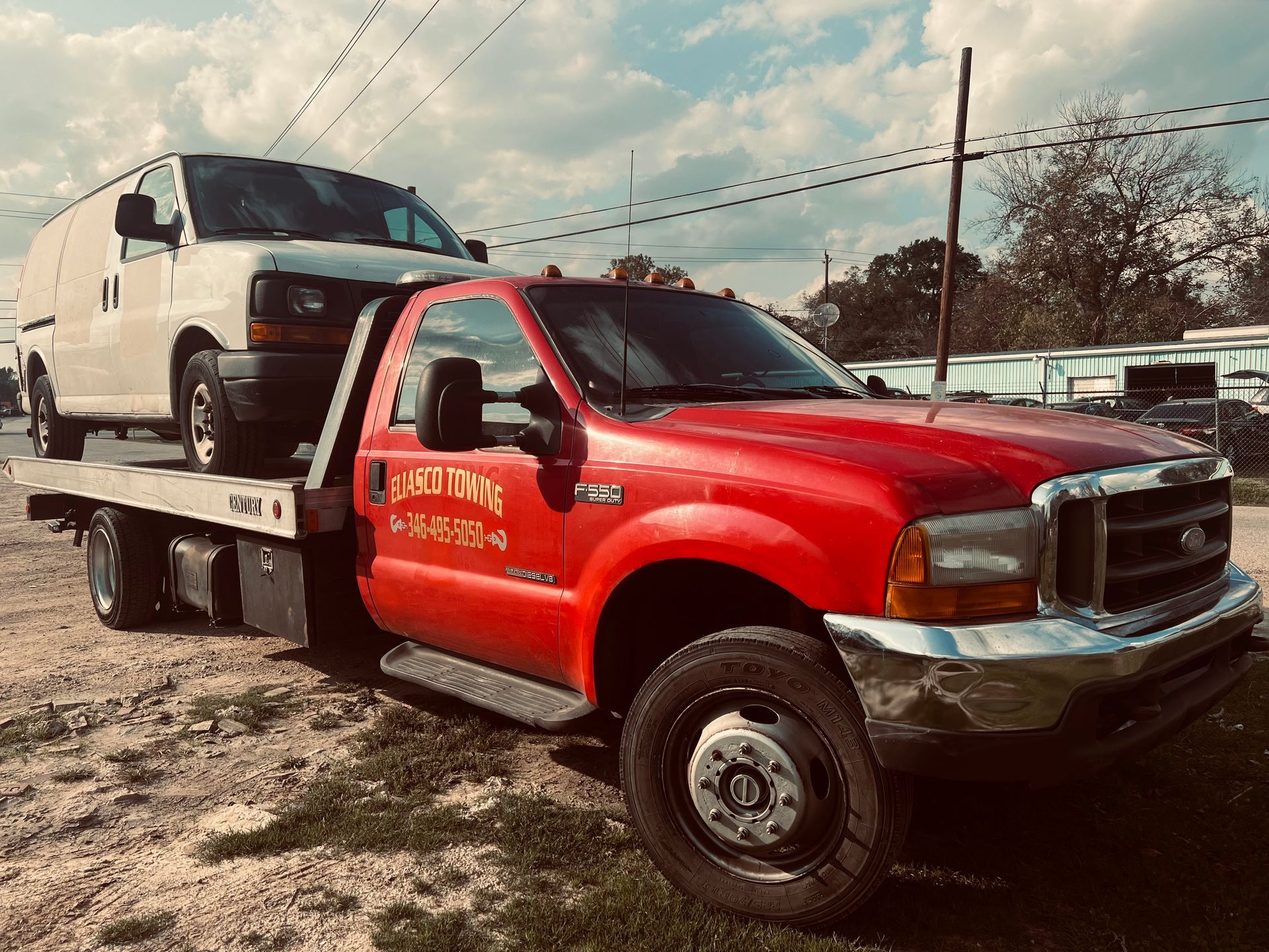 Red tow truck carrying a white SUV in a dirt lot under a cloudy sky