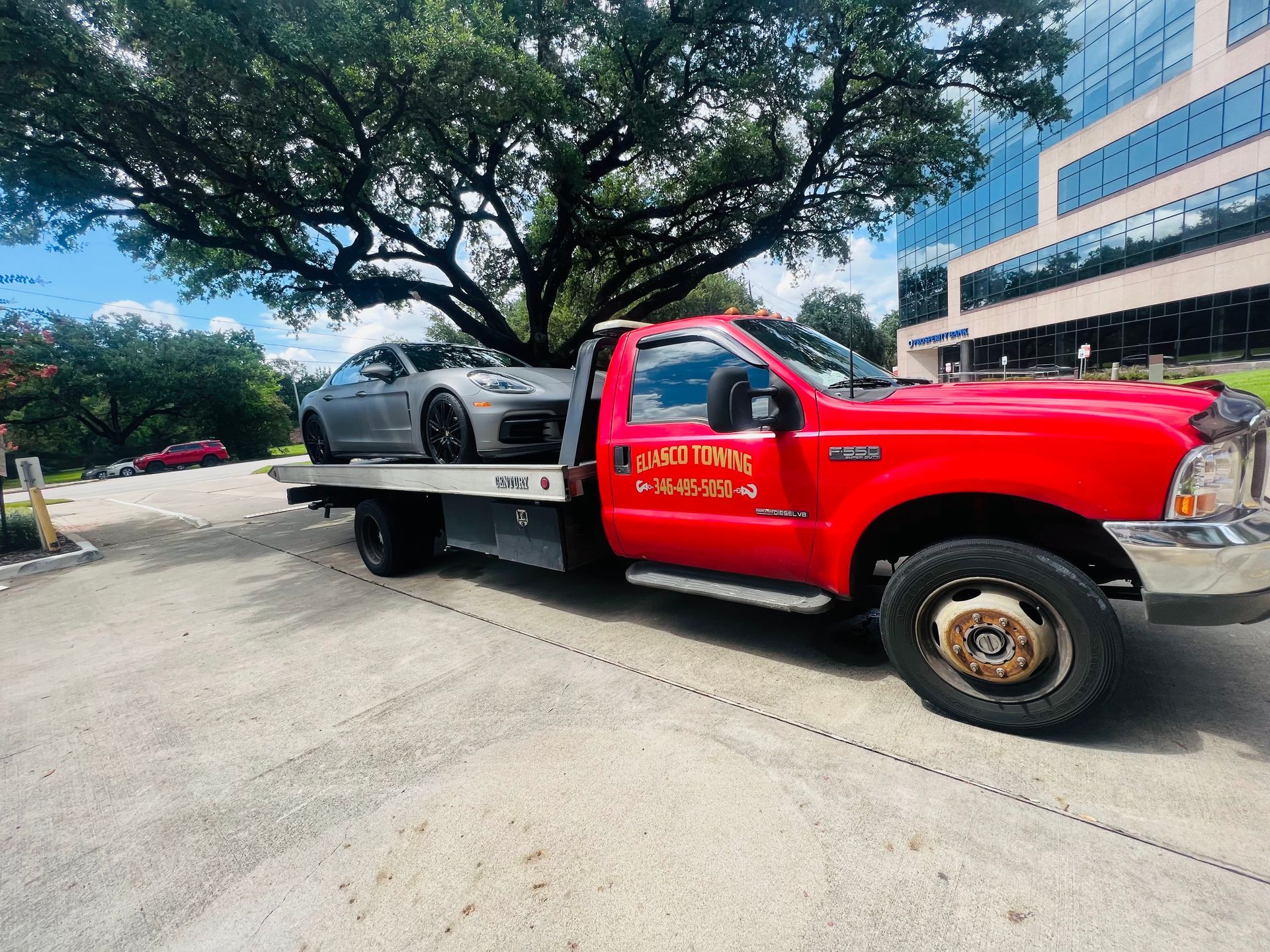 Red tow truck carrying a silver sports car on a street beside a building and trees.