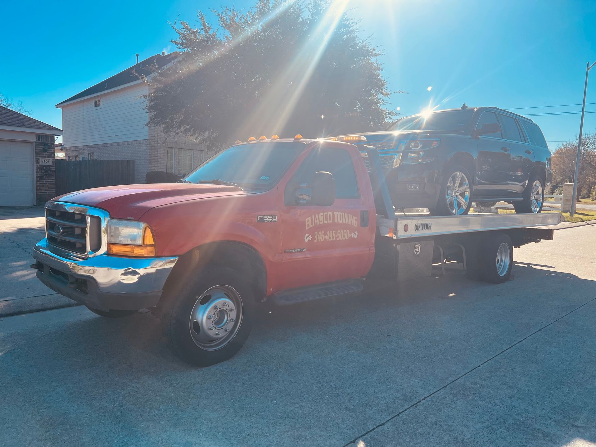 Red flatbed tow truck carrying a black SUV on a sunny street near houses.