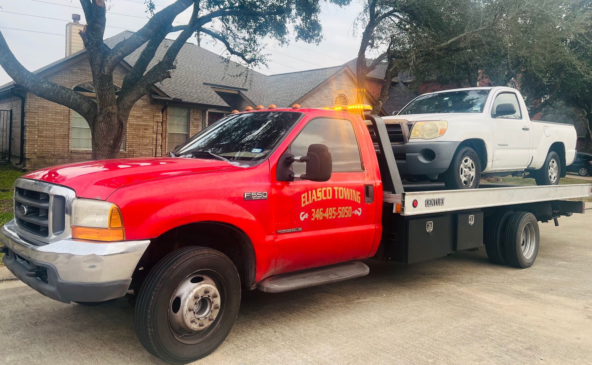 Red tow truck carrying a white pickup on a flatbed in a driveway, amber light flashing.