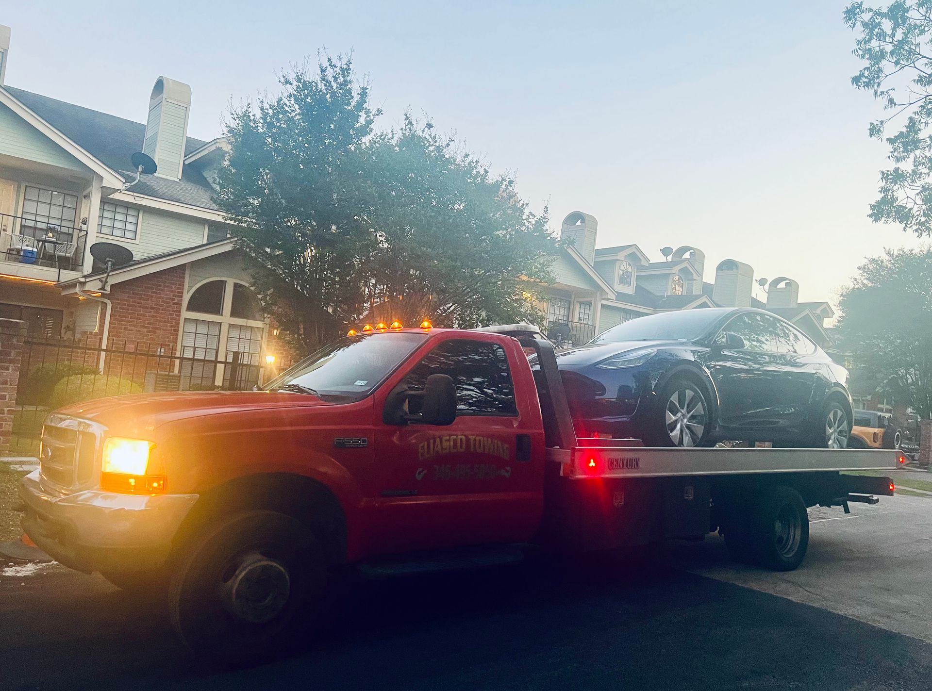 Red tow truck hauling a damaged black SUV on a flatbed at dusk on a residential street