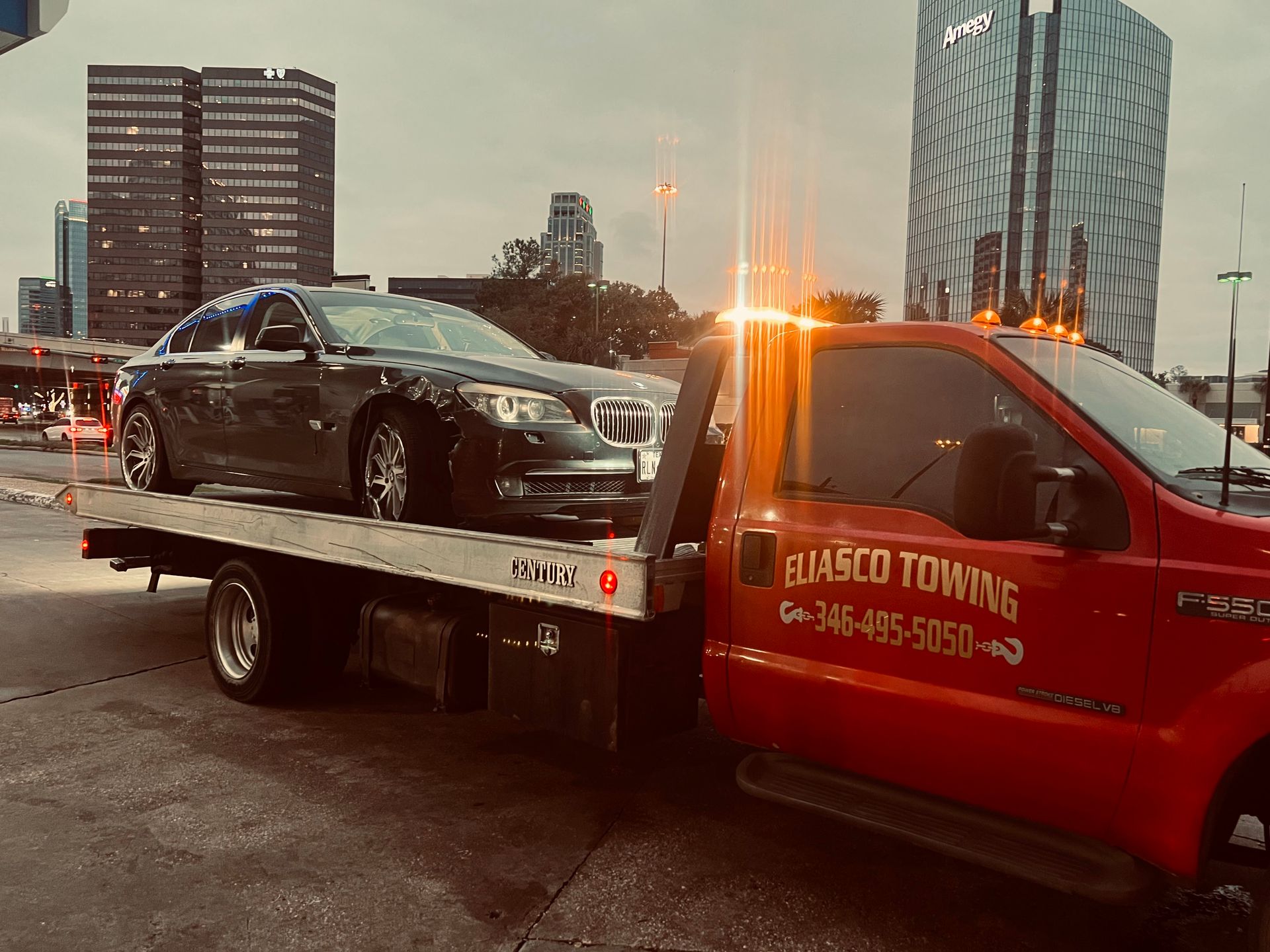 Tow truck hauling a black sedan at dusk in a city street, with a red tow truck and tall buildings nearby