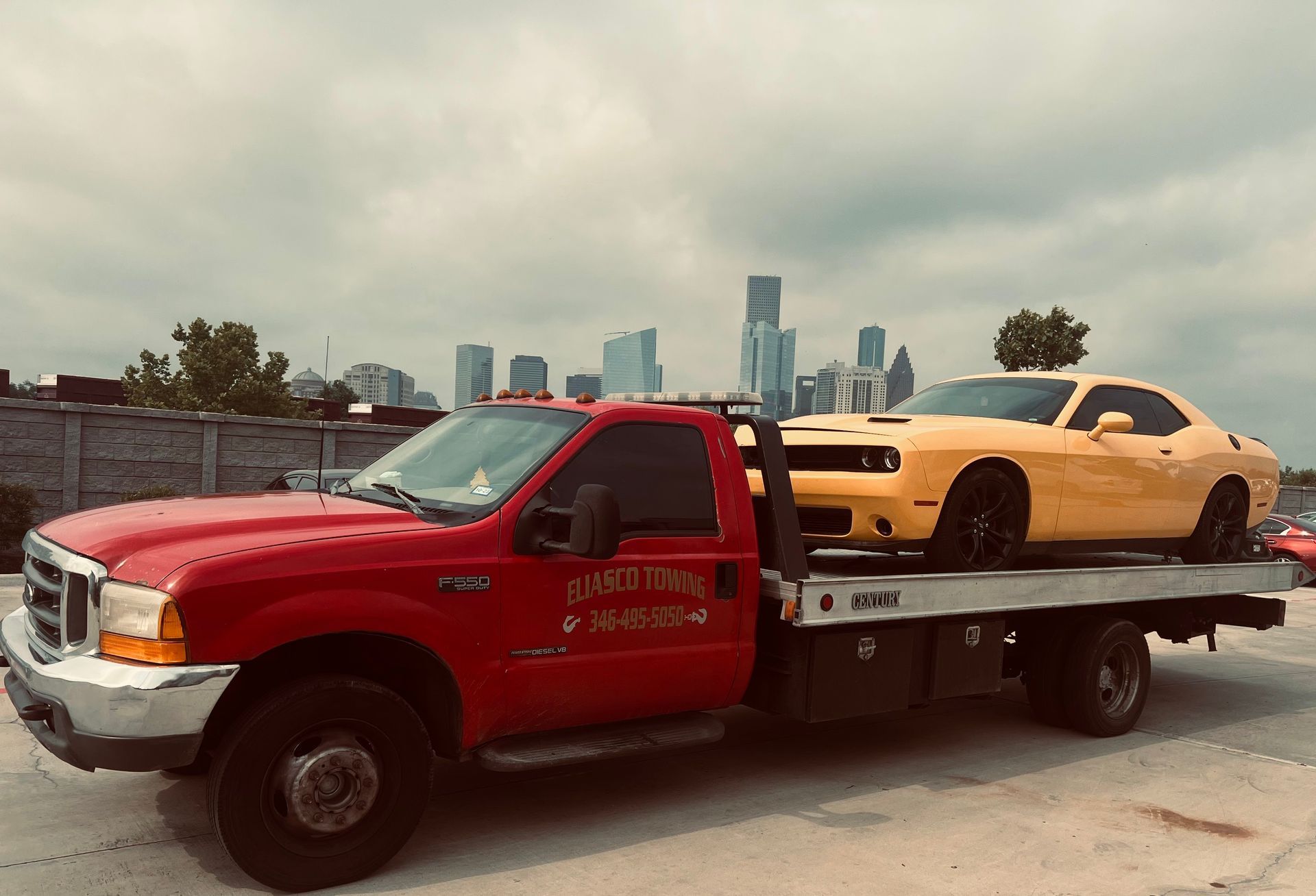 Red tow truck carrying a yellow sports car on a flatbed, with a city skyline in the background.