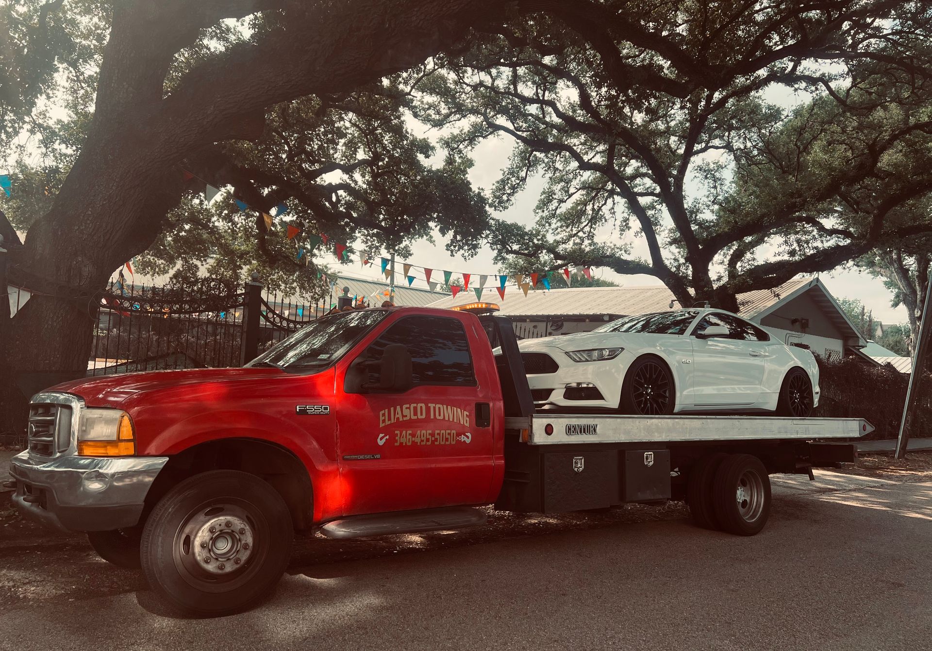 Red tow truck carrying a white car under large trees on a street at dusk