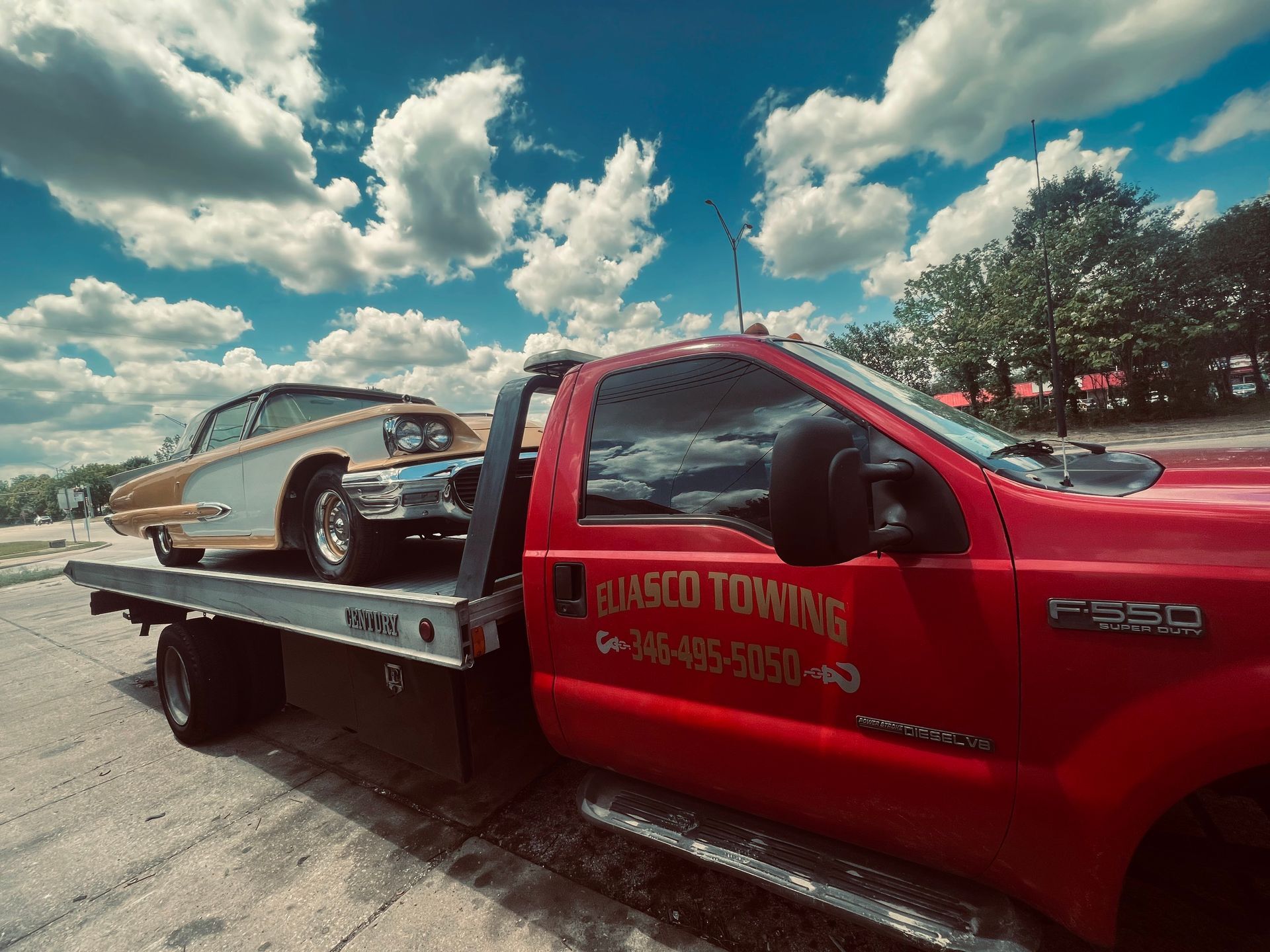 Red tow truck hauling a silver classic car on a flatbed under a bright, cloudy sky.
