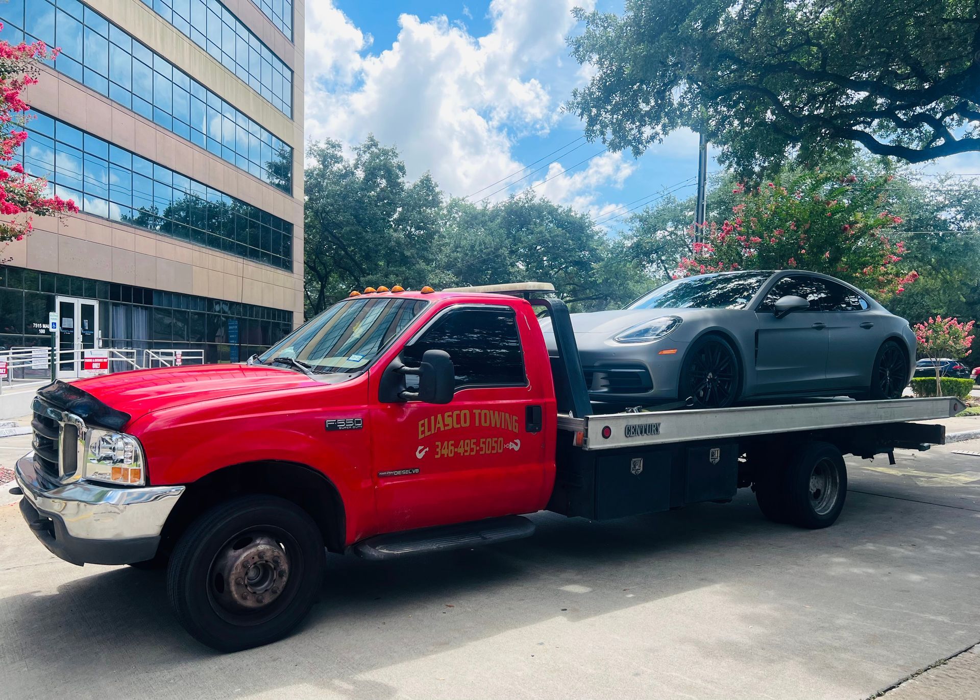 Red tow truck carrying a gray sports car on a city street beside an office building.
