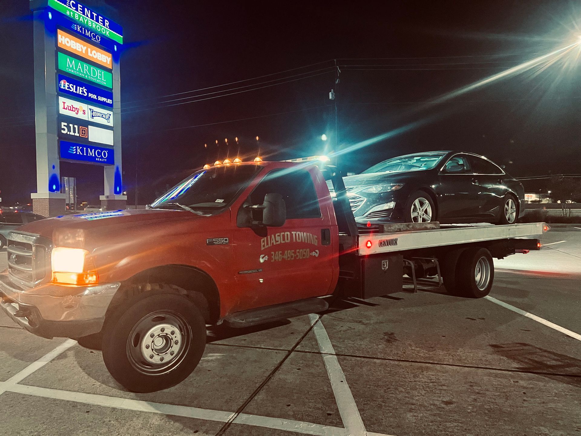 Red tow truck carrying a damaged black car at night in a lit parking lot