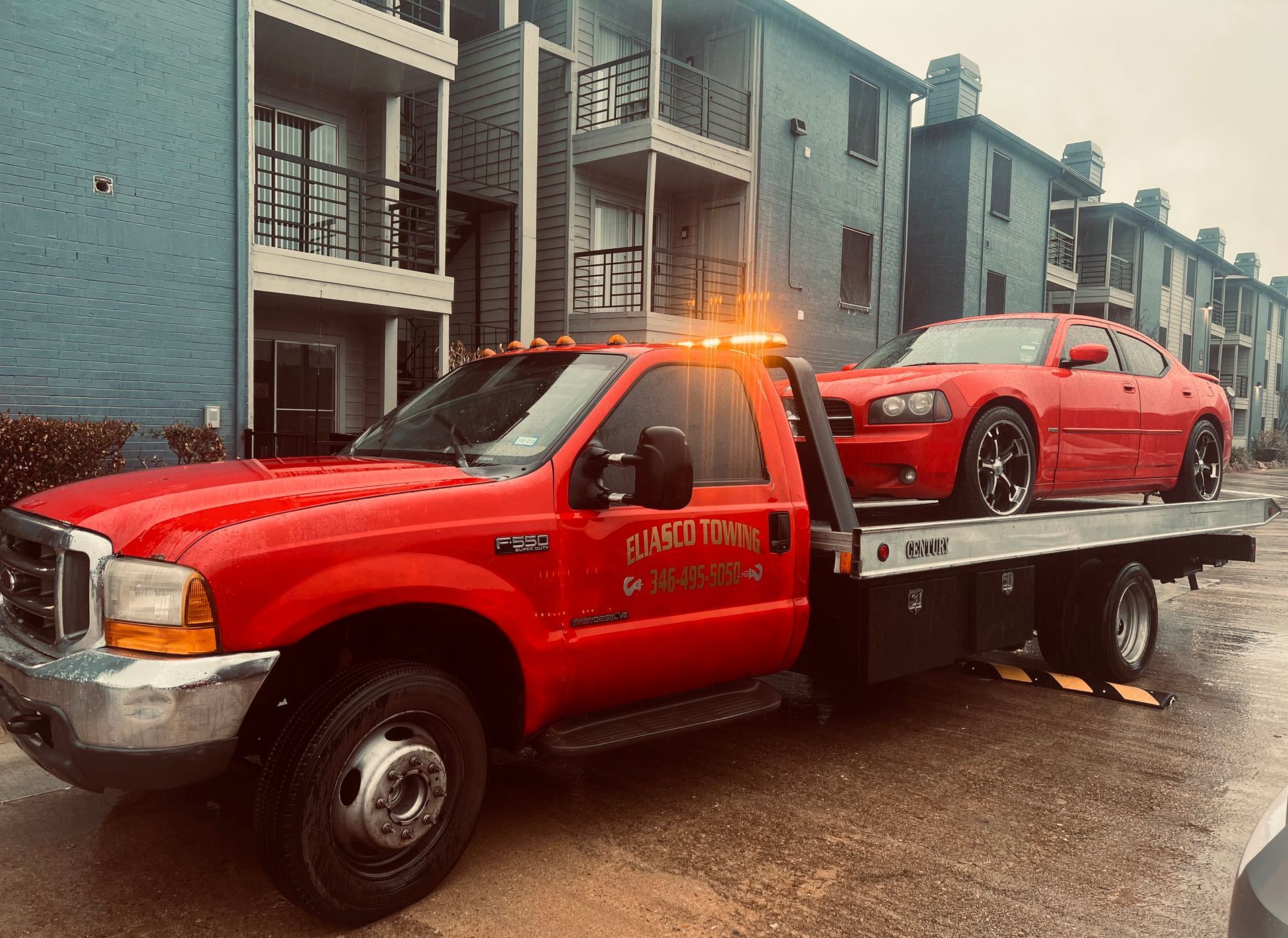 Red tow truck carrying a red sports car on a rainy street beside apartment buildings