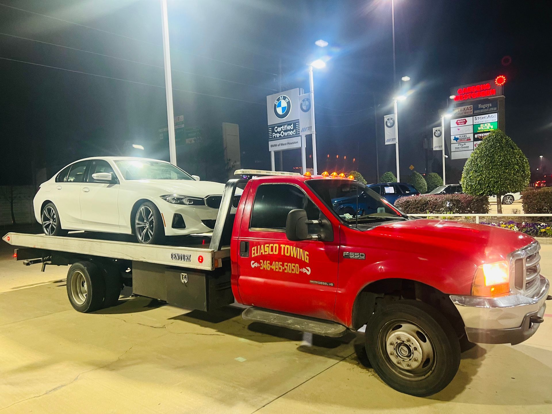 Red tow truck hauling a white BMW on a flatbed at night in a lit parking lot