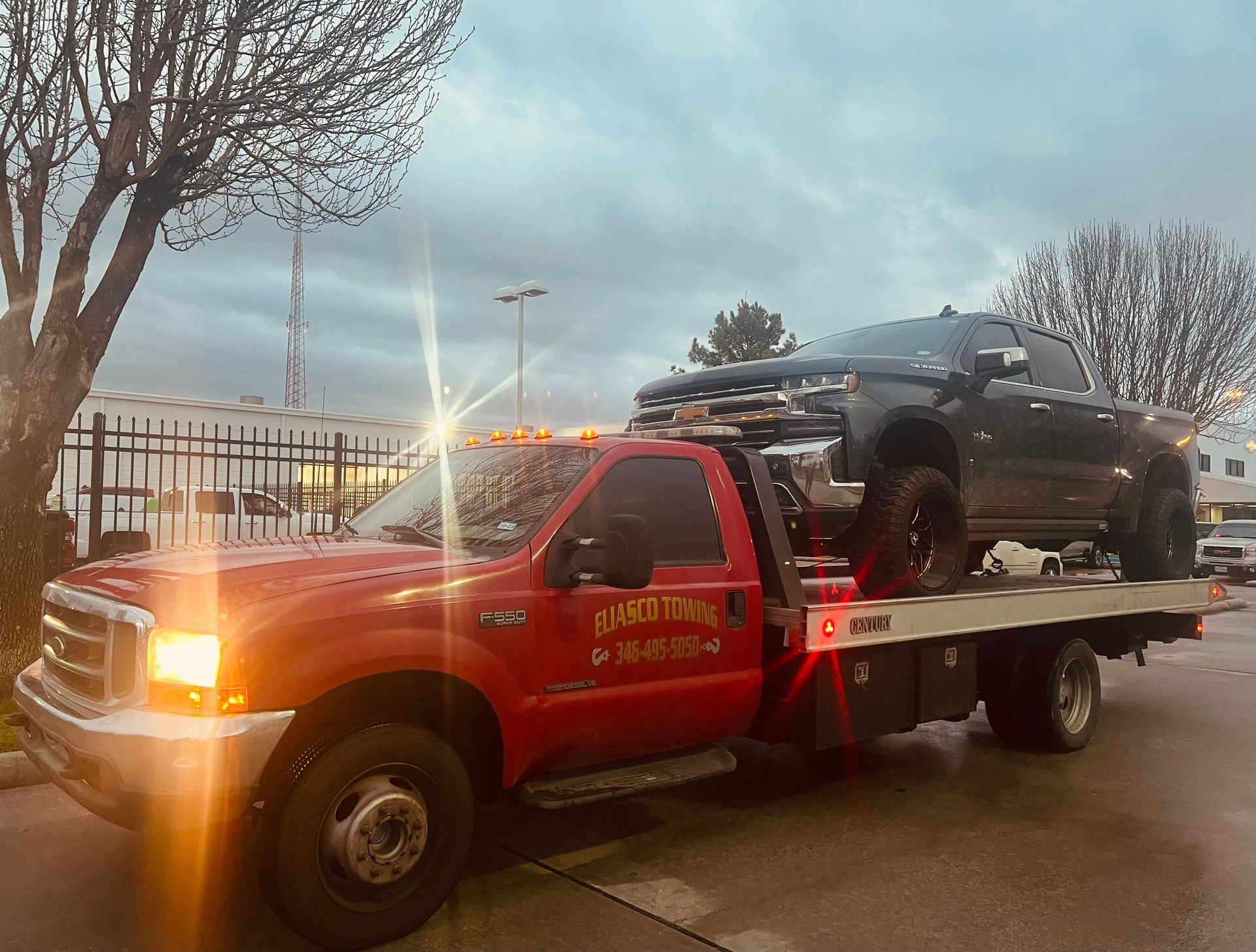 Red tow truck hauling a black SUV on a flatbed at dusk by a fence and trees.