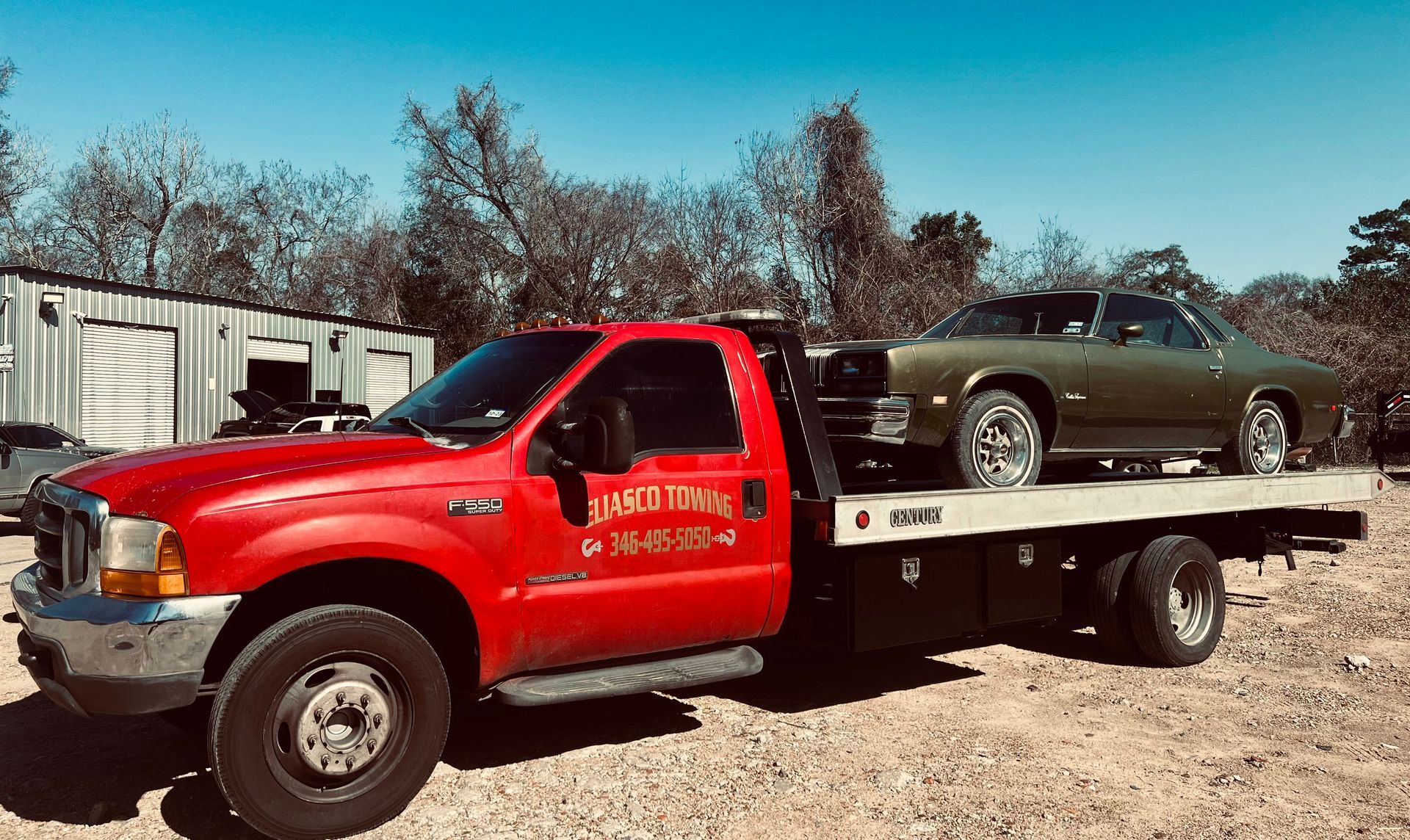 Red tow truck carrying a dark green sedan on a flatbed in a gravel lot