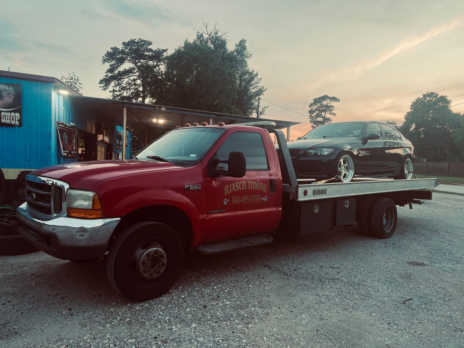 Red tow truck carrying a black car in a gravel lot at dusk