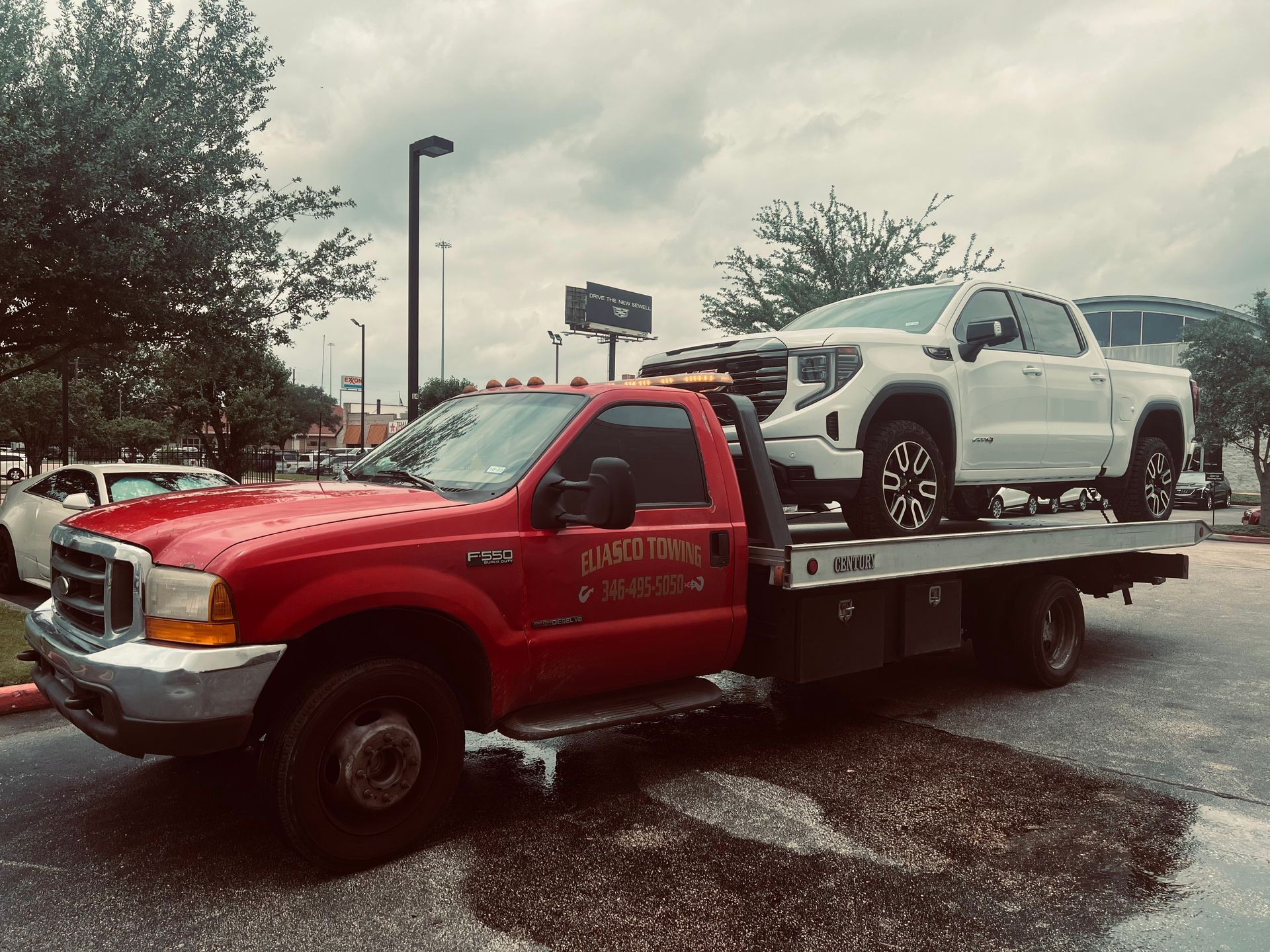 Red tow truck carrying a white pickup on a flatbed in a parking lot during wet weather